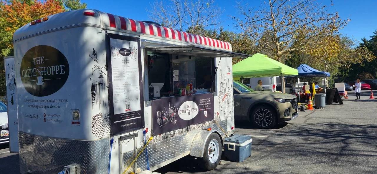 A food truck called 'The Crepe Shoppe' parked at an outdoor market with tents and people, surrounded by trees with fall foliage.