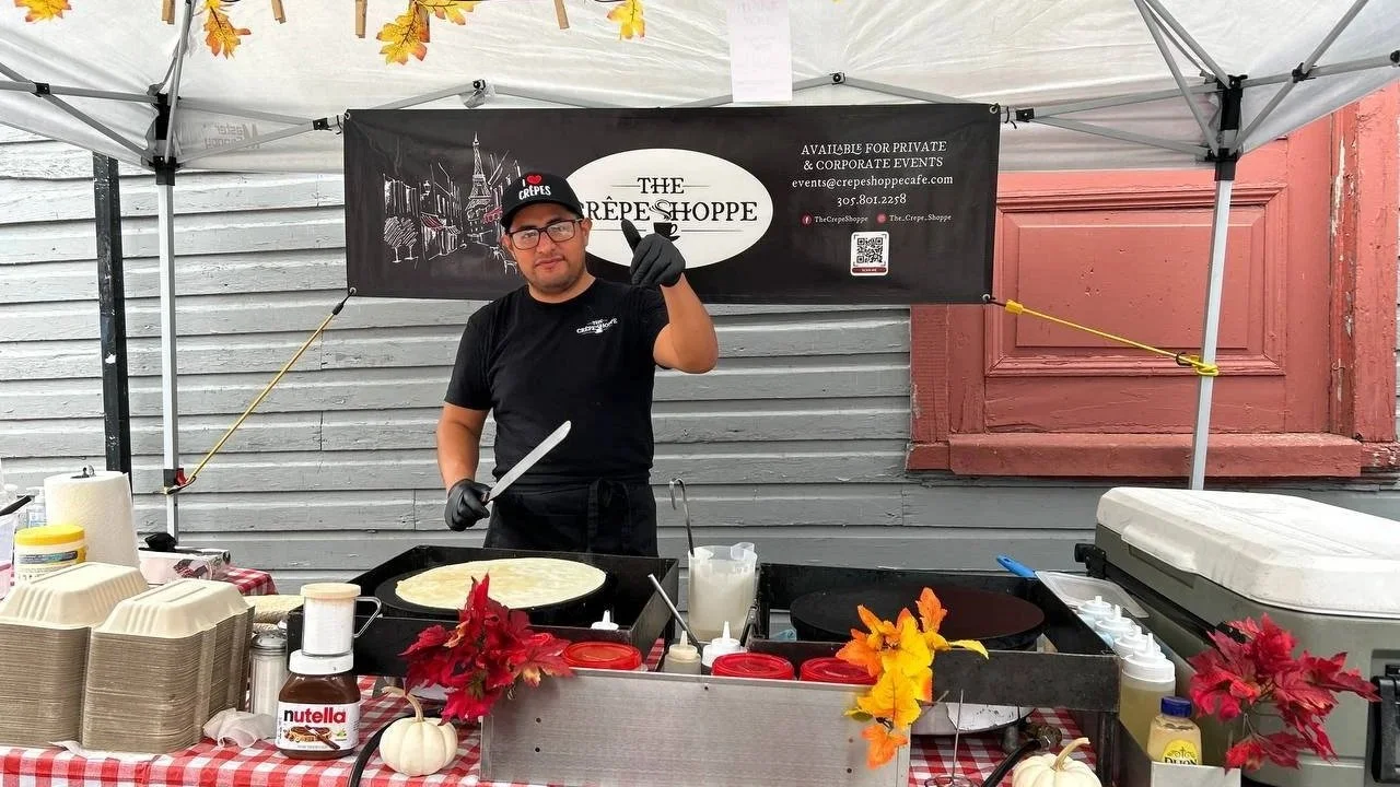Chef at The Crêpe Shoppe Café making fresh dessert crêpes on a heated pan at an outdoor private wedding catering setup in Annapolis, wearing black cap, glasses, and gloves, with fall leaves and pumpkins decorating the mobile crepe station and branded