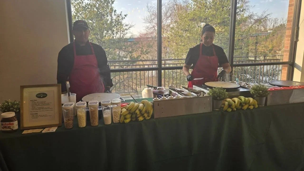 Chef at The Crêpe Shoppe Café preparing fresh crêpes indoors at a corporate staff appreciation week catering event, wearing a black cap, glasses, black t-shirt and gloves, standing behind the mobile crepe station with fall leaves and pumpkins as deco