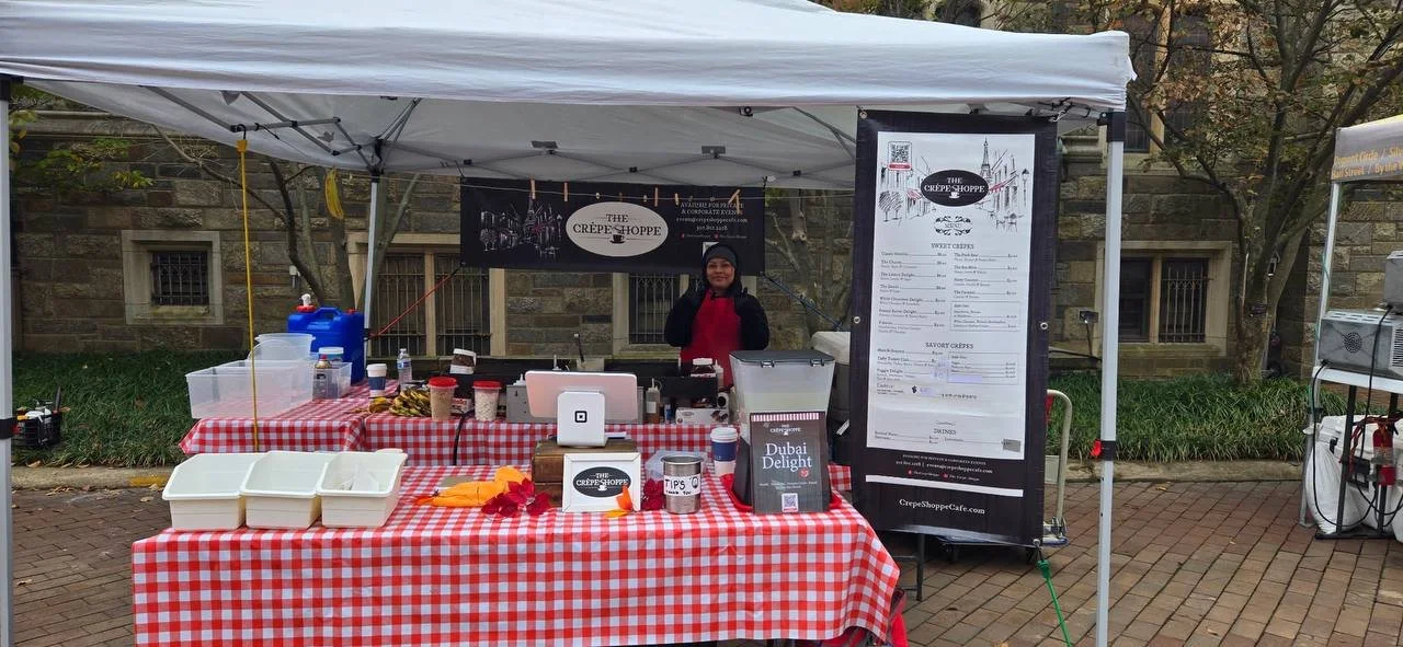 A person standing behind a food stall with a red and white checkered tablecloth, serving crepes. The stall has a menu and tips jar, with a tent canopy above and trees in the background.