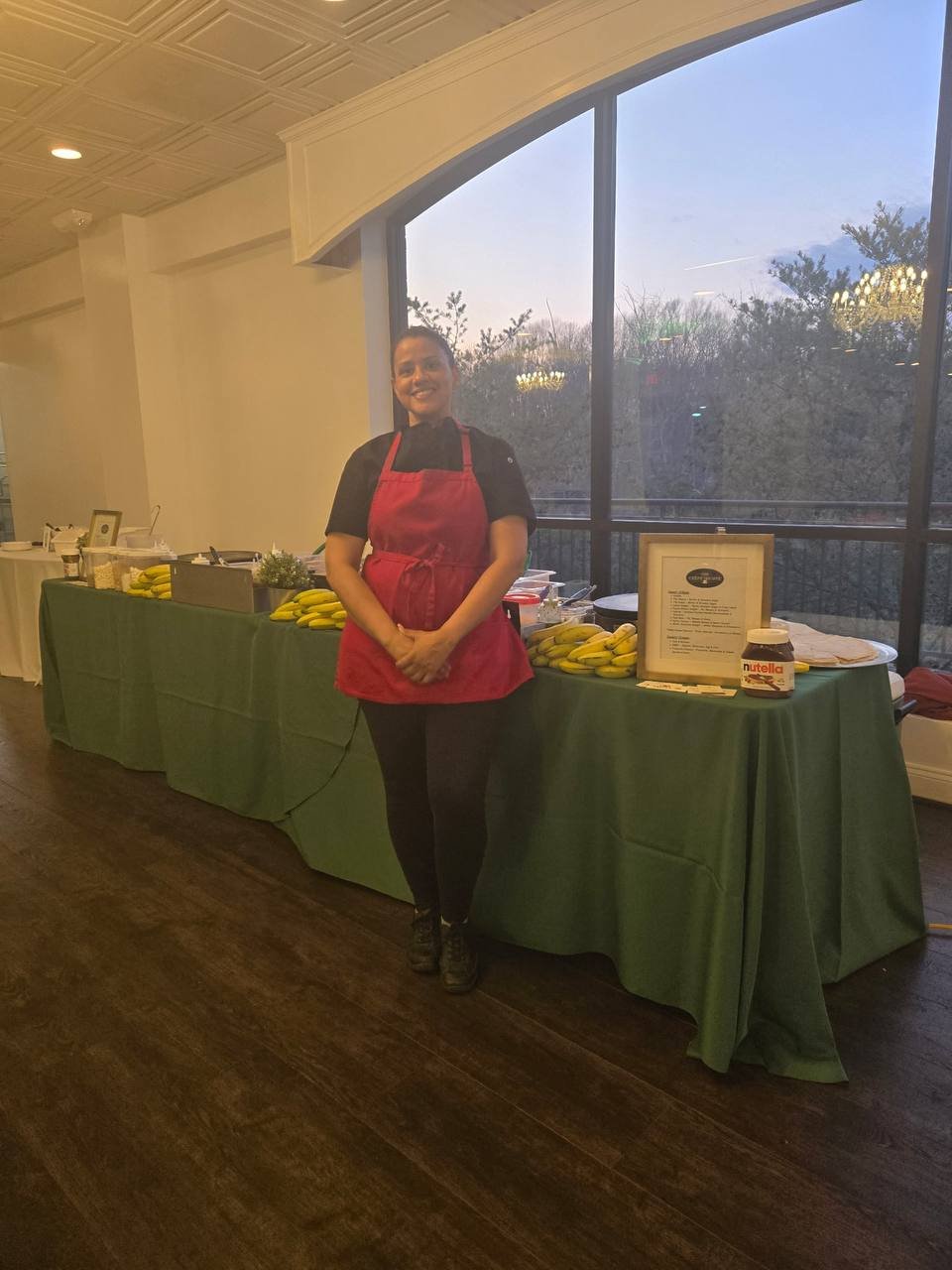 A smiling woman wearing a red apron stands next to a green-covered table filled with bananas, a Nutella jar, and framed menu, inside a room with large windows showing trees outside during the evening.