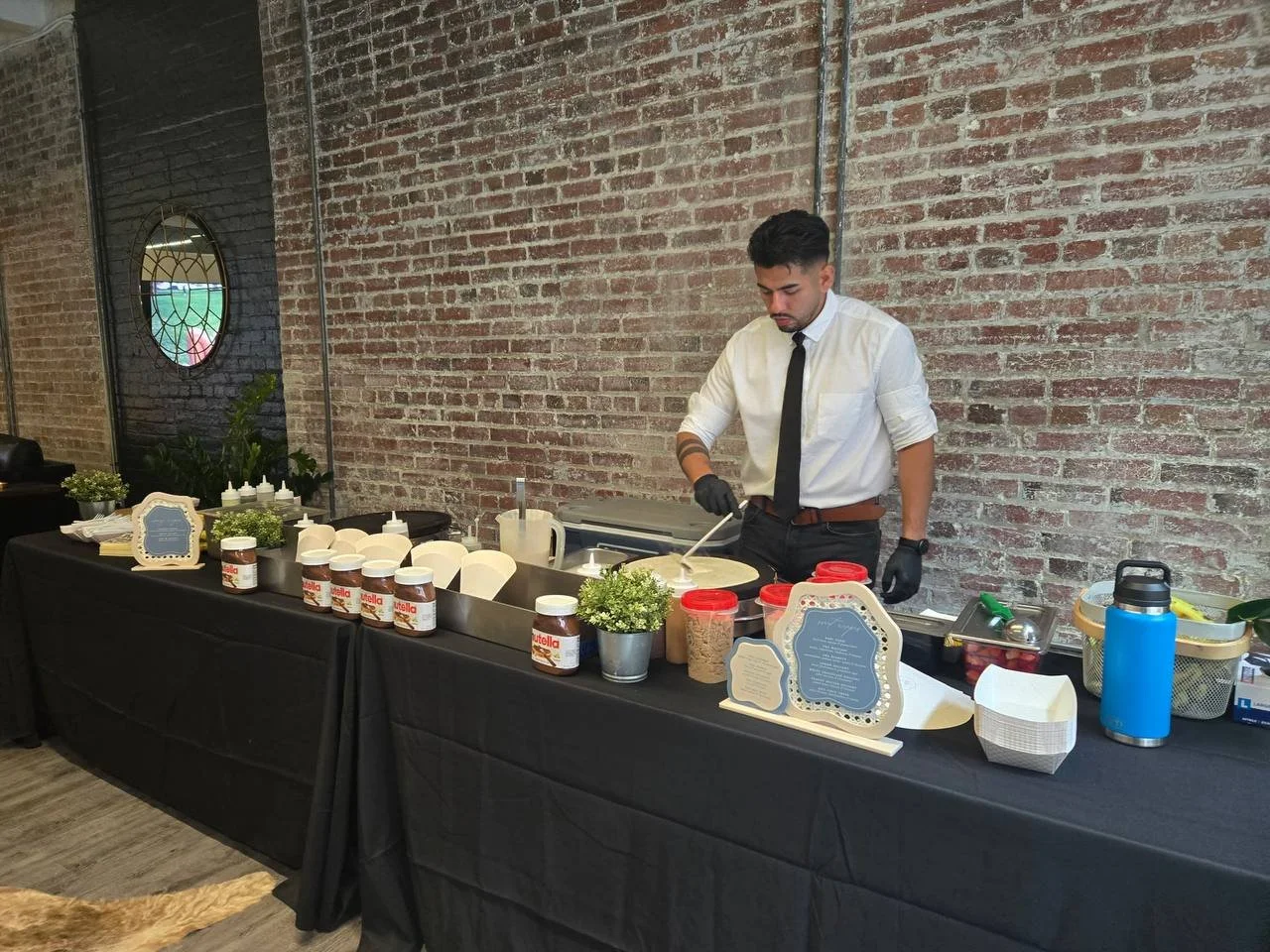 A man in a white shirt, black tie, and black gloves is making crepes at a dessert station with jars of Nutella, bowls of toppings, and potted plants on a black table against an exposed brick wall.
