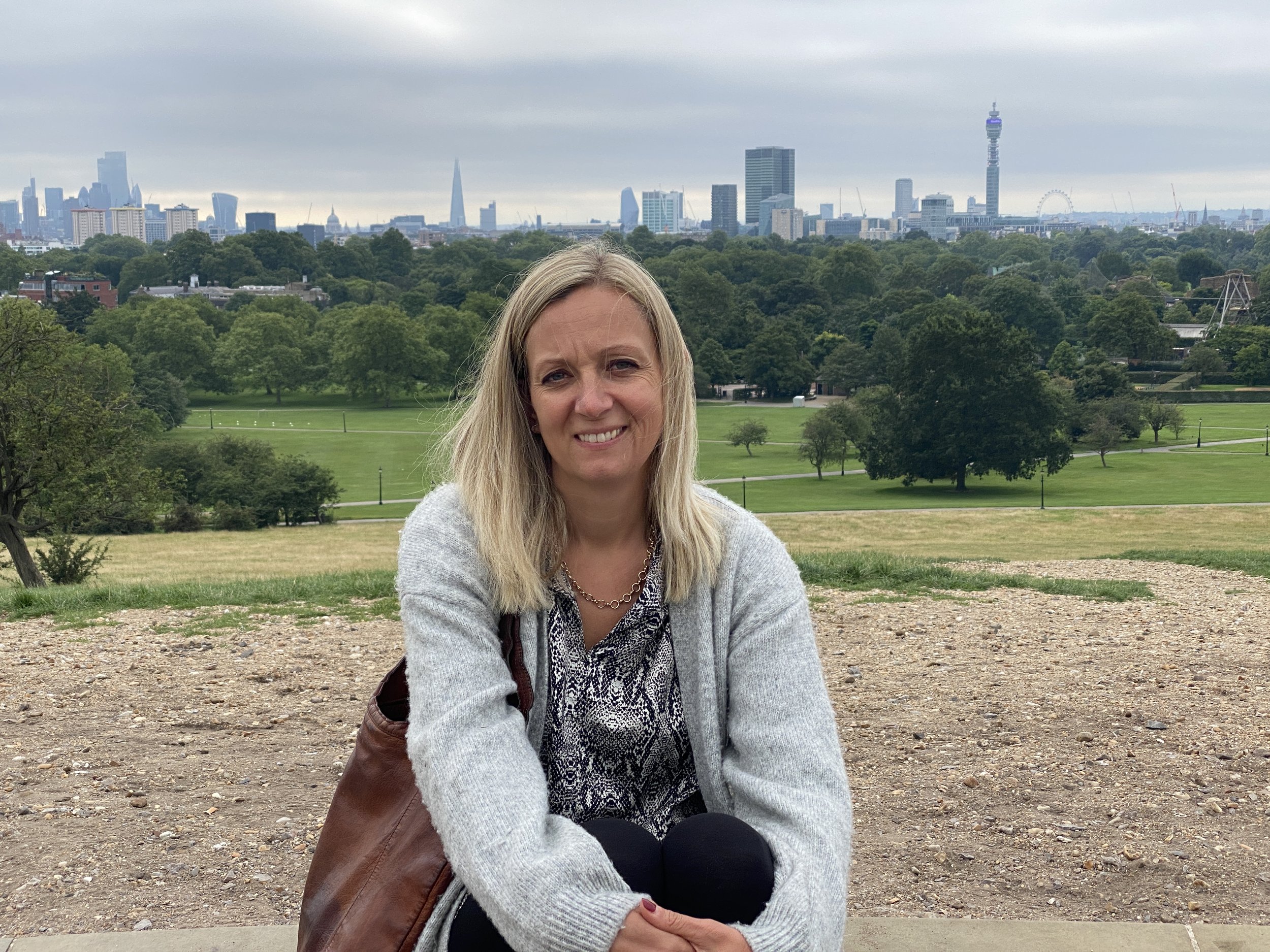 A woman with blonde hair sitting outdoors on a gravel area in a park with a city skyline in the background. There are trees and grassy areas in the park. The weather is overcast.