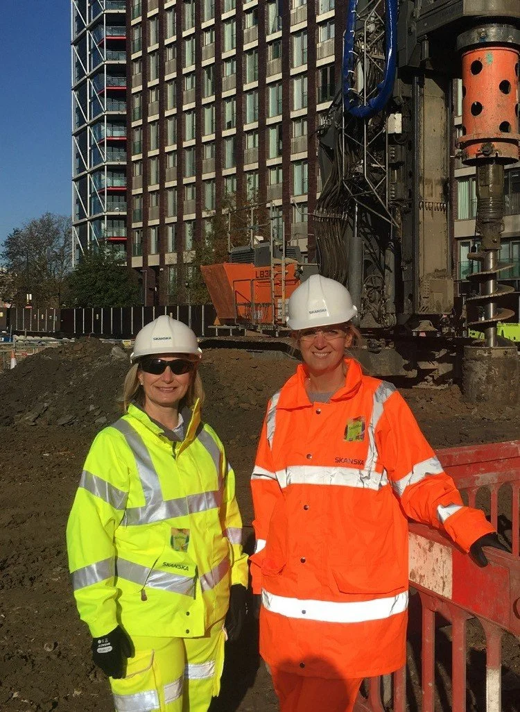Two female construction workers in safety helmets and reflective jackets, standing on a construction site with a large building in the background.