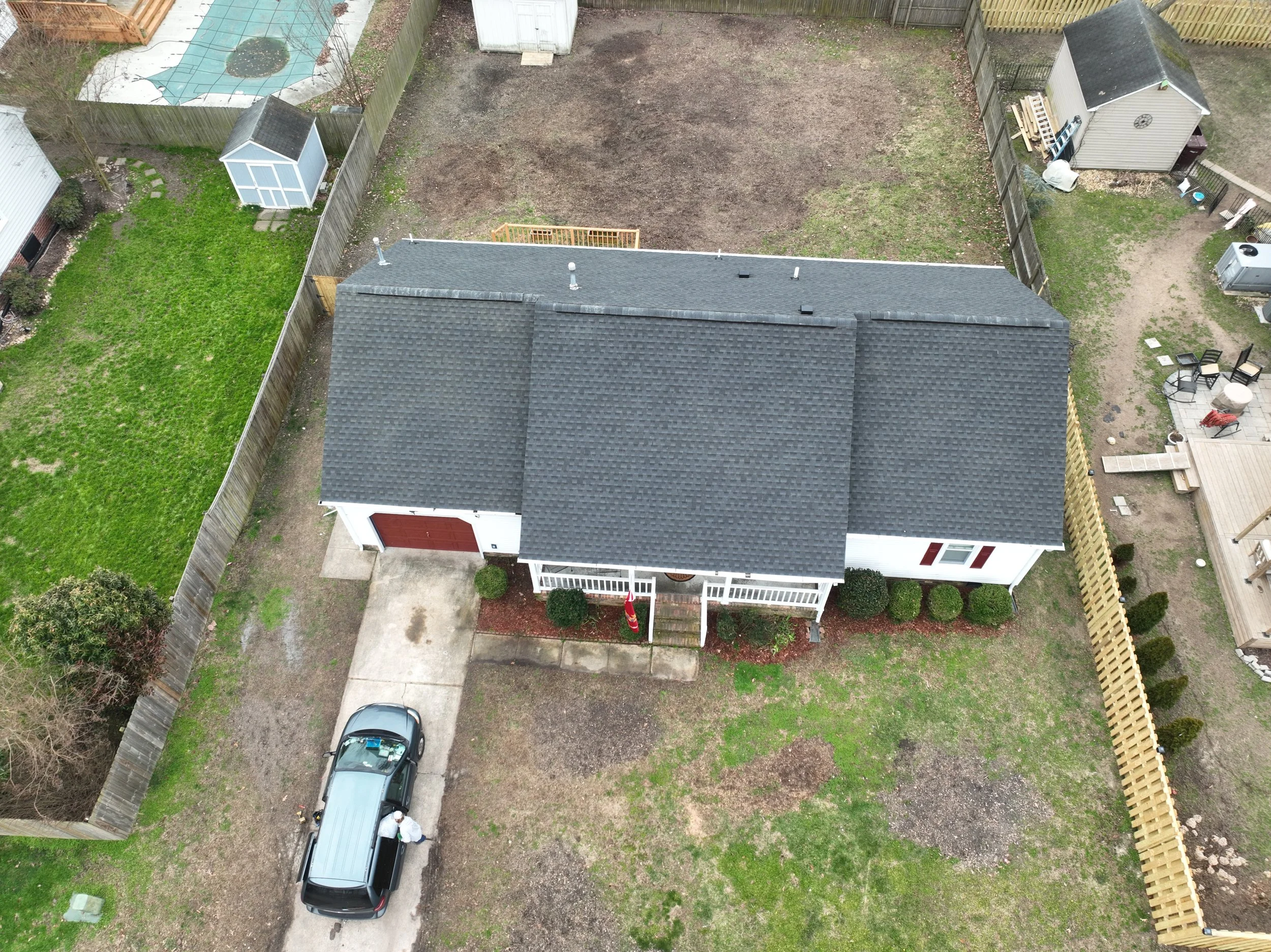 An aerial view of a residential backyard showing a house with a gray shingled roof, a driveway with a black car, a grassy lawn, a small shed, a fenced garden area, and an uncovered dirt area likely for a future project.