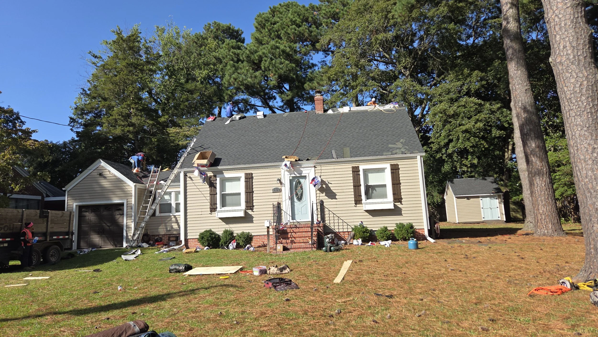 Workers repairing the roof of a two-story house on a sunny day, with tools and materials scattered on the lawn and a ladder leaning against the house.