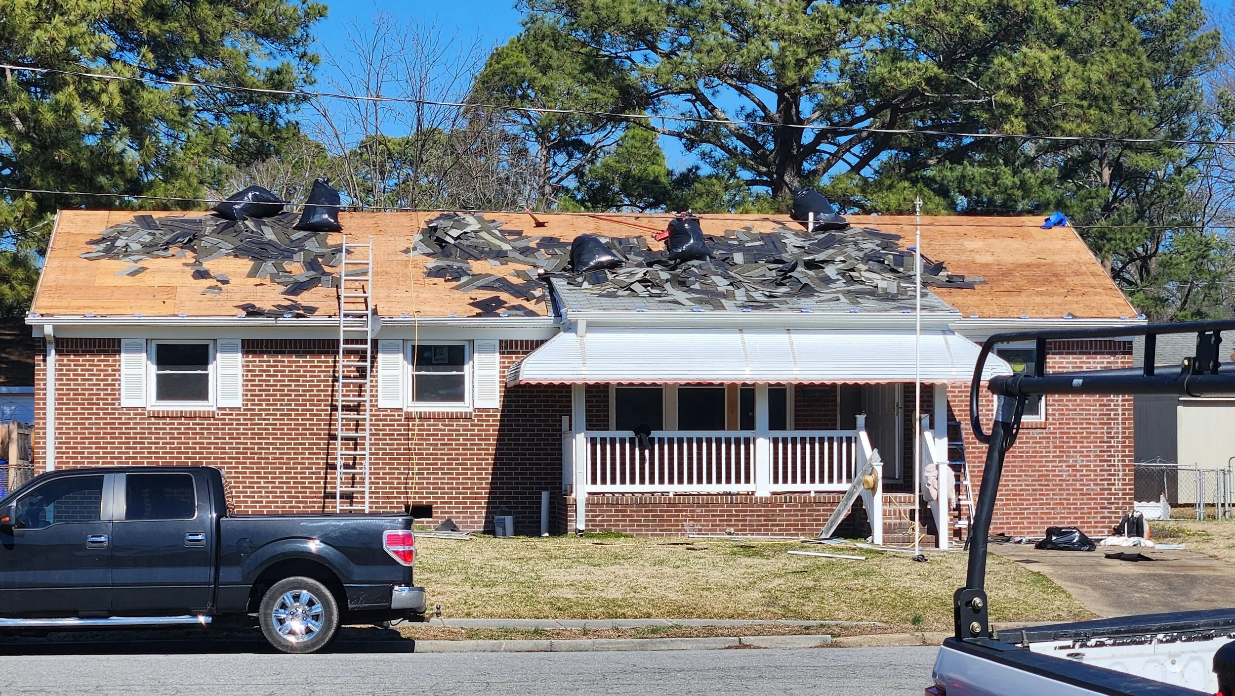 Residential house under renovation with a new roof being installed, construction materials and tools on the roof, a black truck parked in front, and trees in the background.