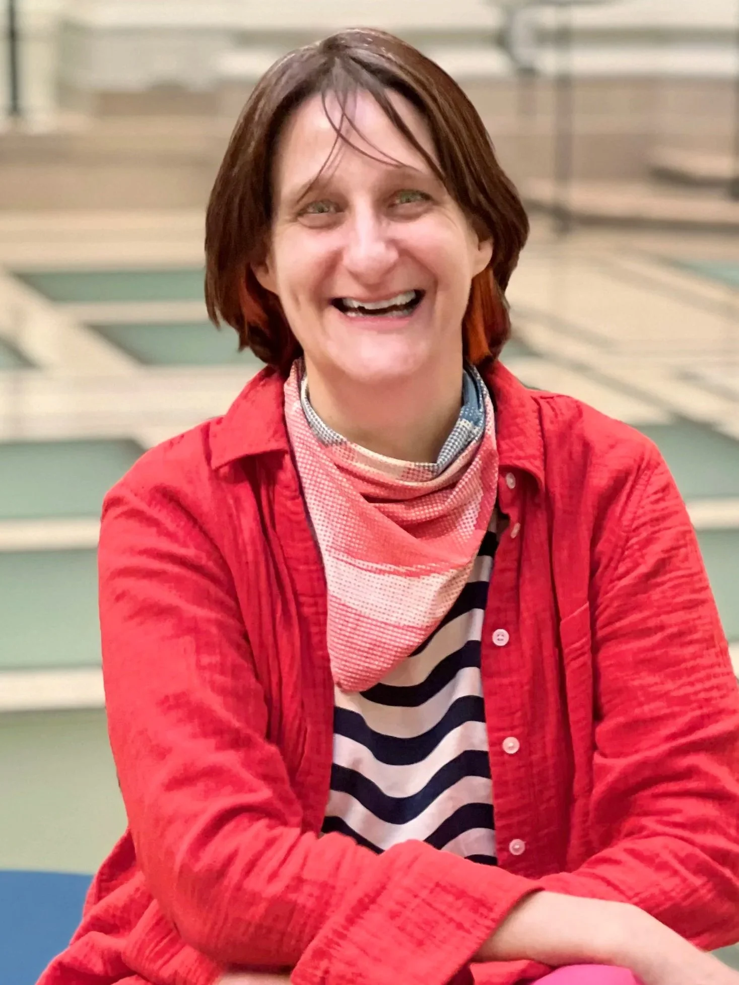 A woman with short brown hair, smiling and wearing a red jacket over a striped shirt, sitting outdoors in front of a set of stairs.
