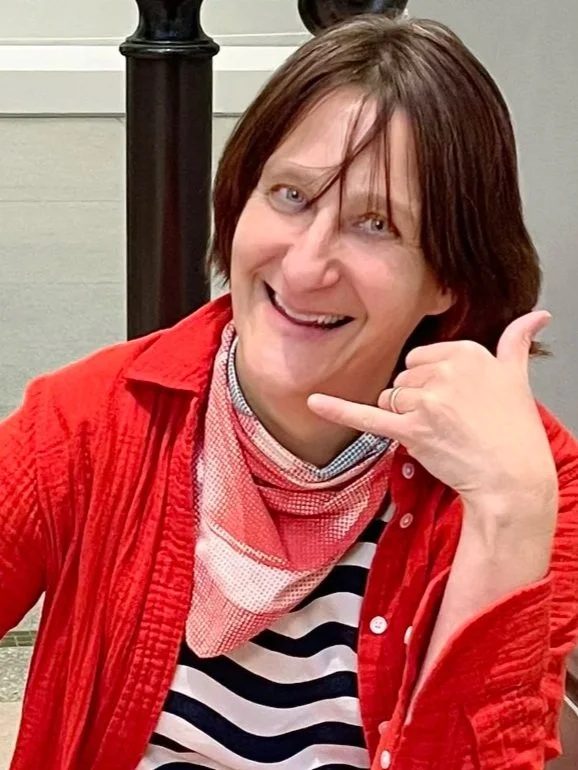 A smiling woman with short brown hair, wearing a red jacket over a black and white striped shirt, making a phone gesture with her hand near her face.