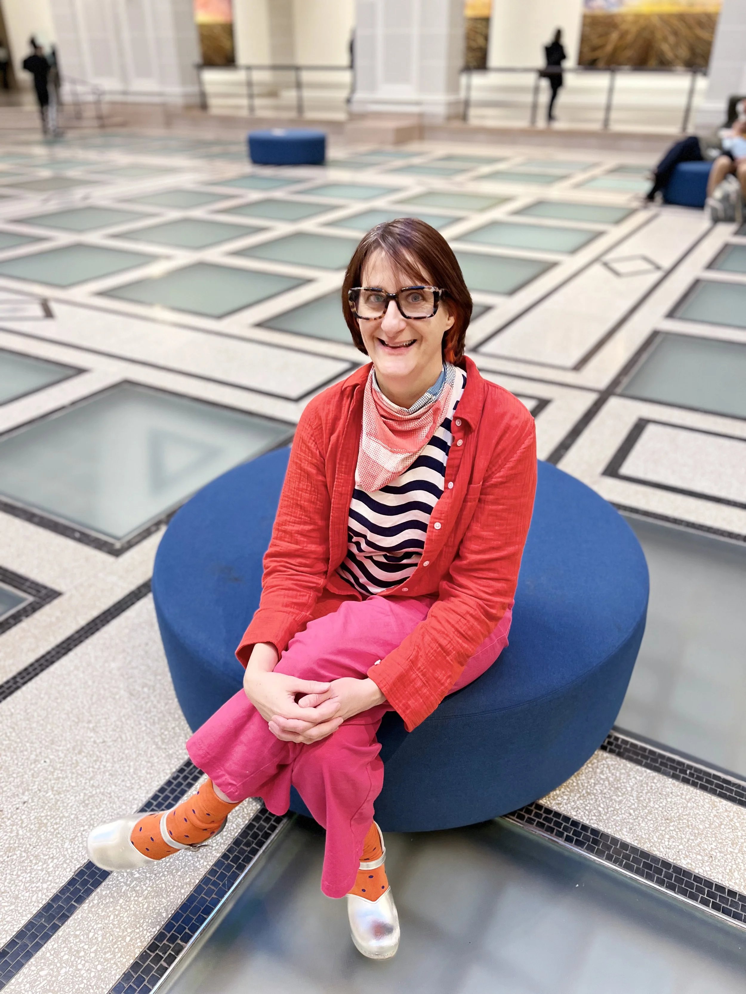 A woman sitting on a round blue seat, smiling, wearing glasses, a red shirt over a striped blue and white shirt, pink pants, colorful socks, and metallic shoes, in a spacious indoor area with geometric tile flooring.