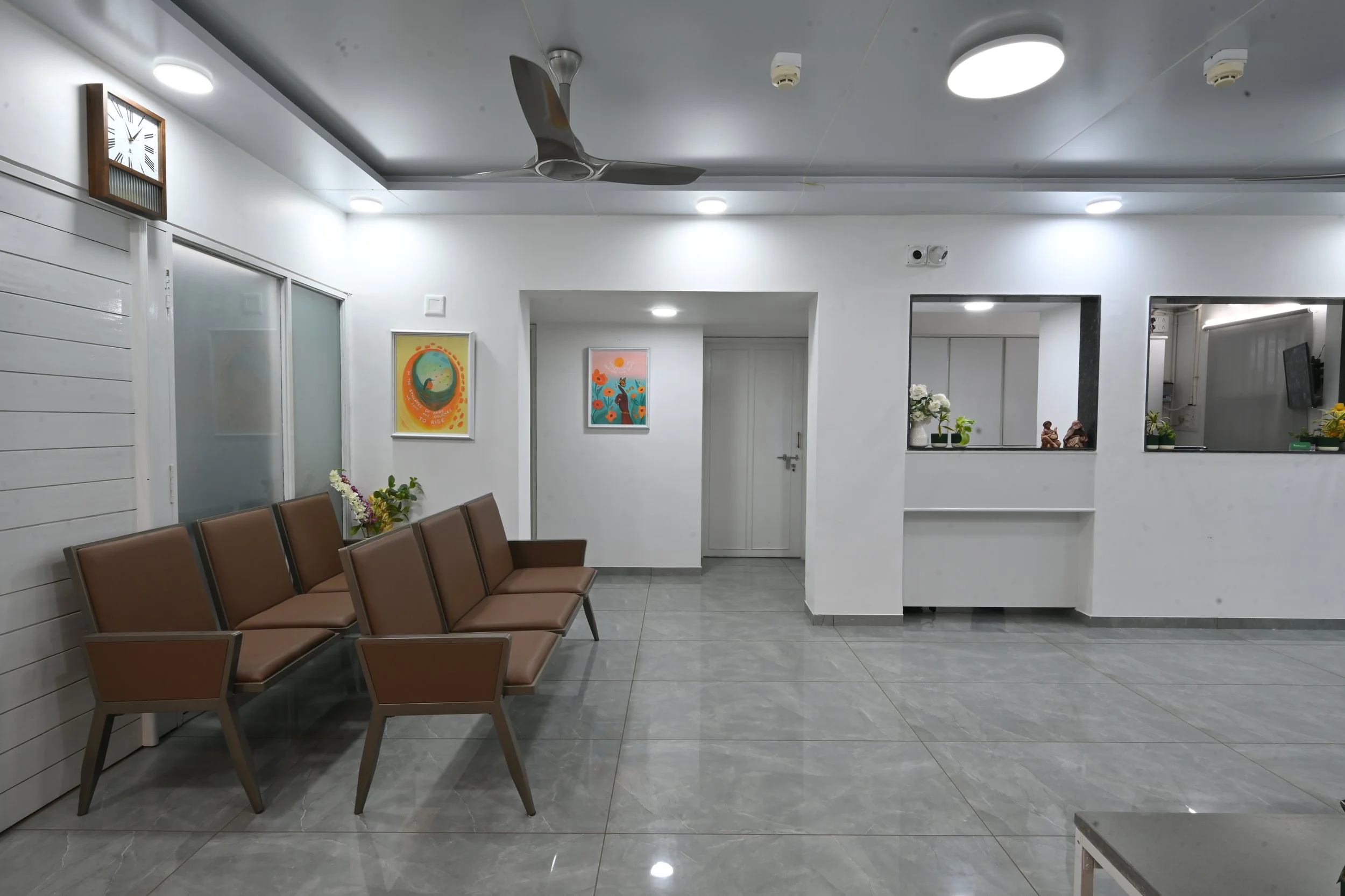 Empty waiting room with brown chairs, white walls, colorful framed pictures, flowers, and a large clock. There is a window counter and ceiling fans.