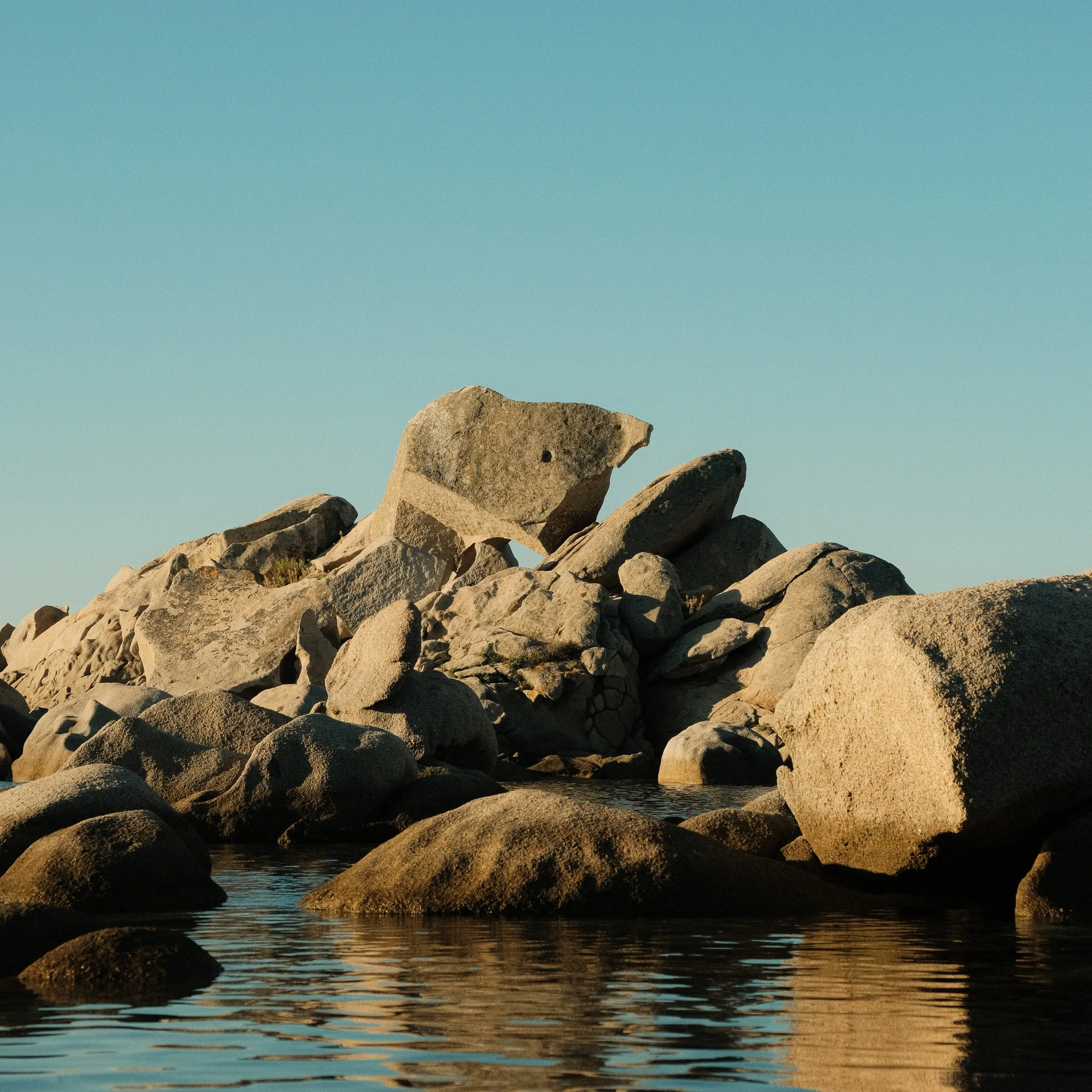 Gruppe von großen, glatten Steinen am Ufer eines ruhigen Gewässers unter einem klaren blauen Himmel.