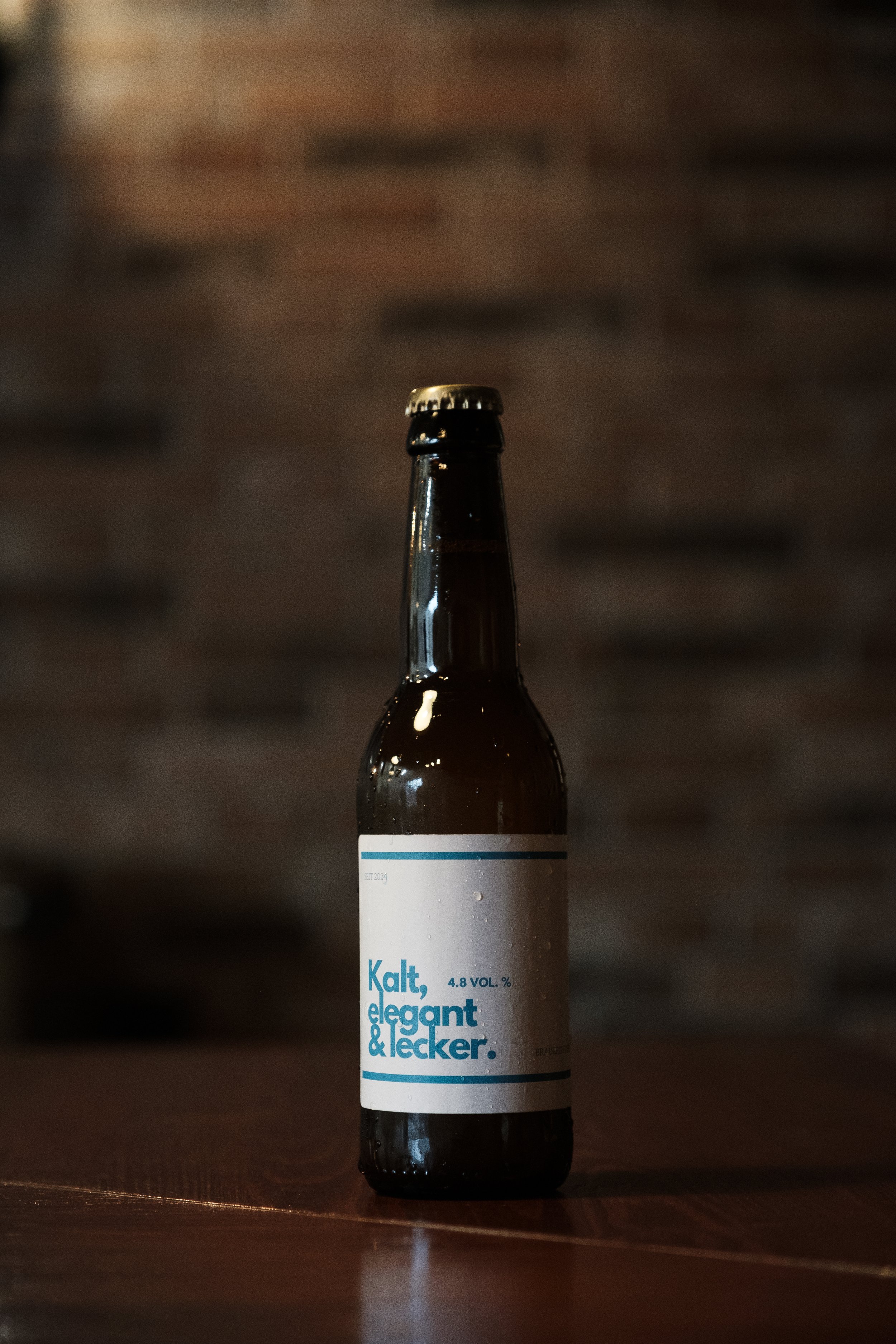 Eine dunkle Flasche Bier mit Weißem Etikett auf einem Holztisch, Hintergrund aus dunklen Ziegeln.