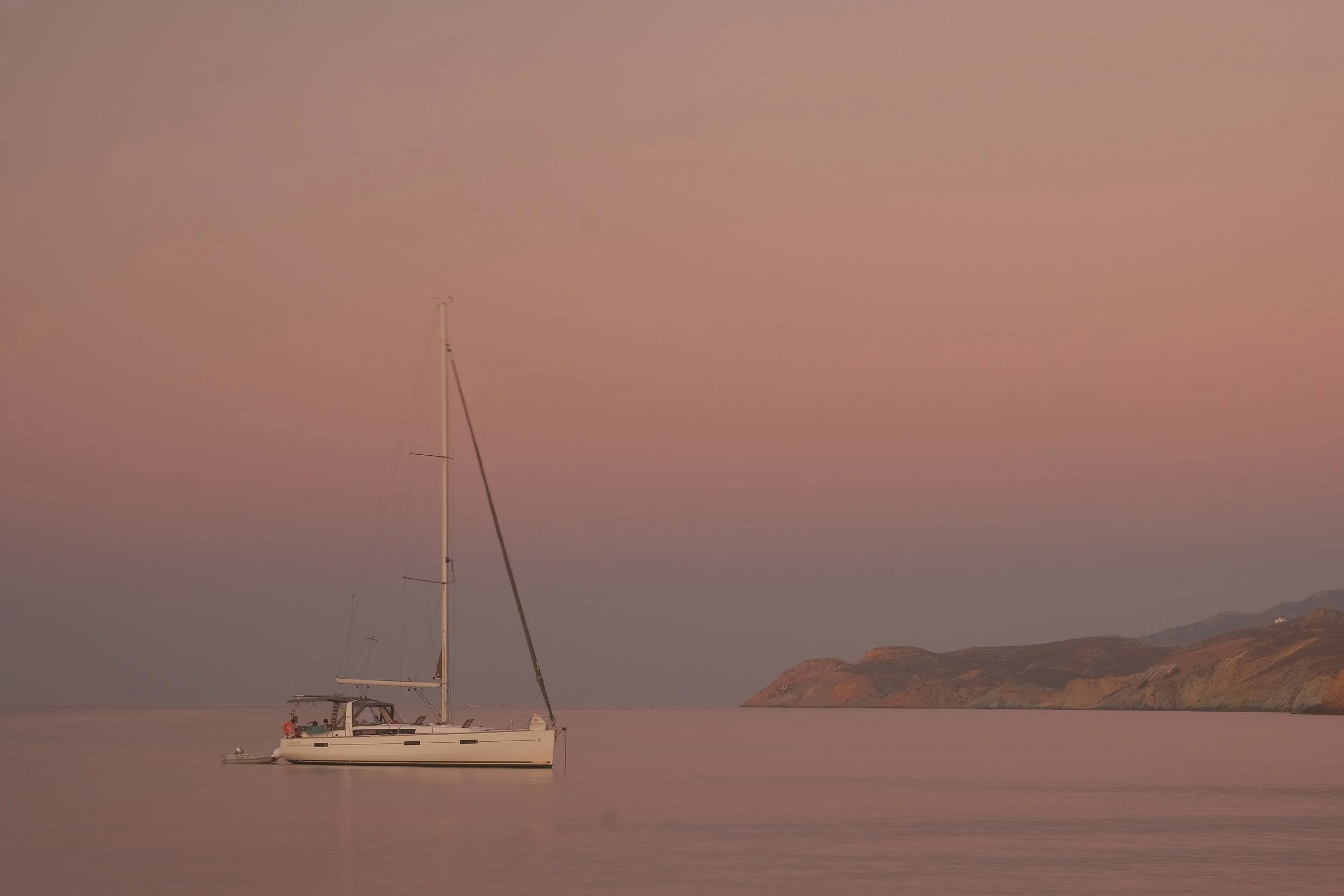 Segelboot auf ruhigem Wasser bei Sonnenuntergang mit rosafarbenem Himmel und Bergen im Hintergrund.