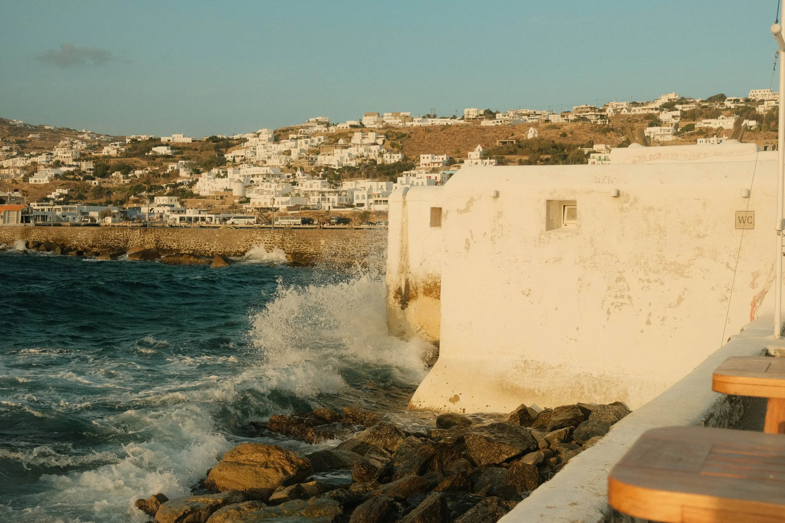 Meeresküste in Santorini, Griechenland, mit weißen Gebäuden auf Hügeln im Hintergrund, Wasser schäumt gegen die Felsen, Sonnenlicht.