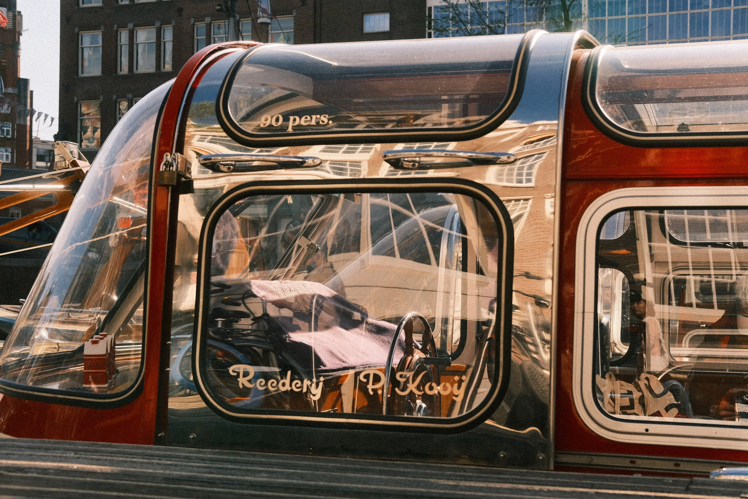 Rote und transparente Fußgängerfähre mit Innenraum, Fahrradsitz und einem pinken Handtuch, mit reflektierenden Gebäuden im Hintergrund.