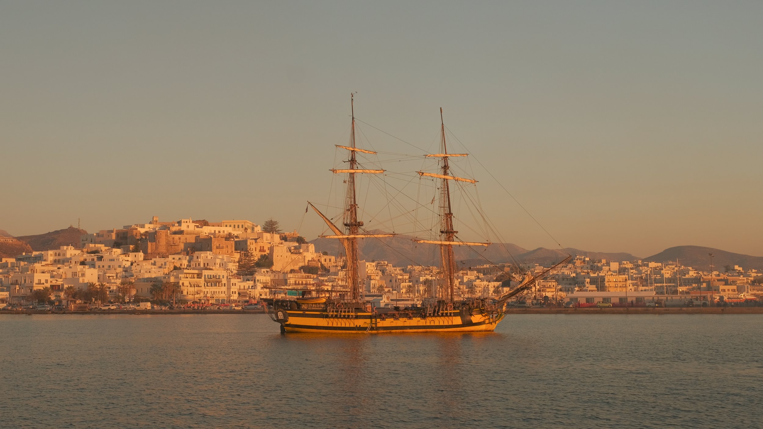 Historisches Segelschiff vor einer Küste mit weißen Gebäuden bei Sonnenuntergang in Griechenland