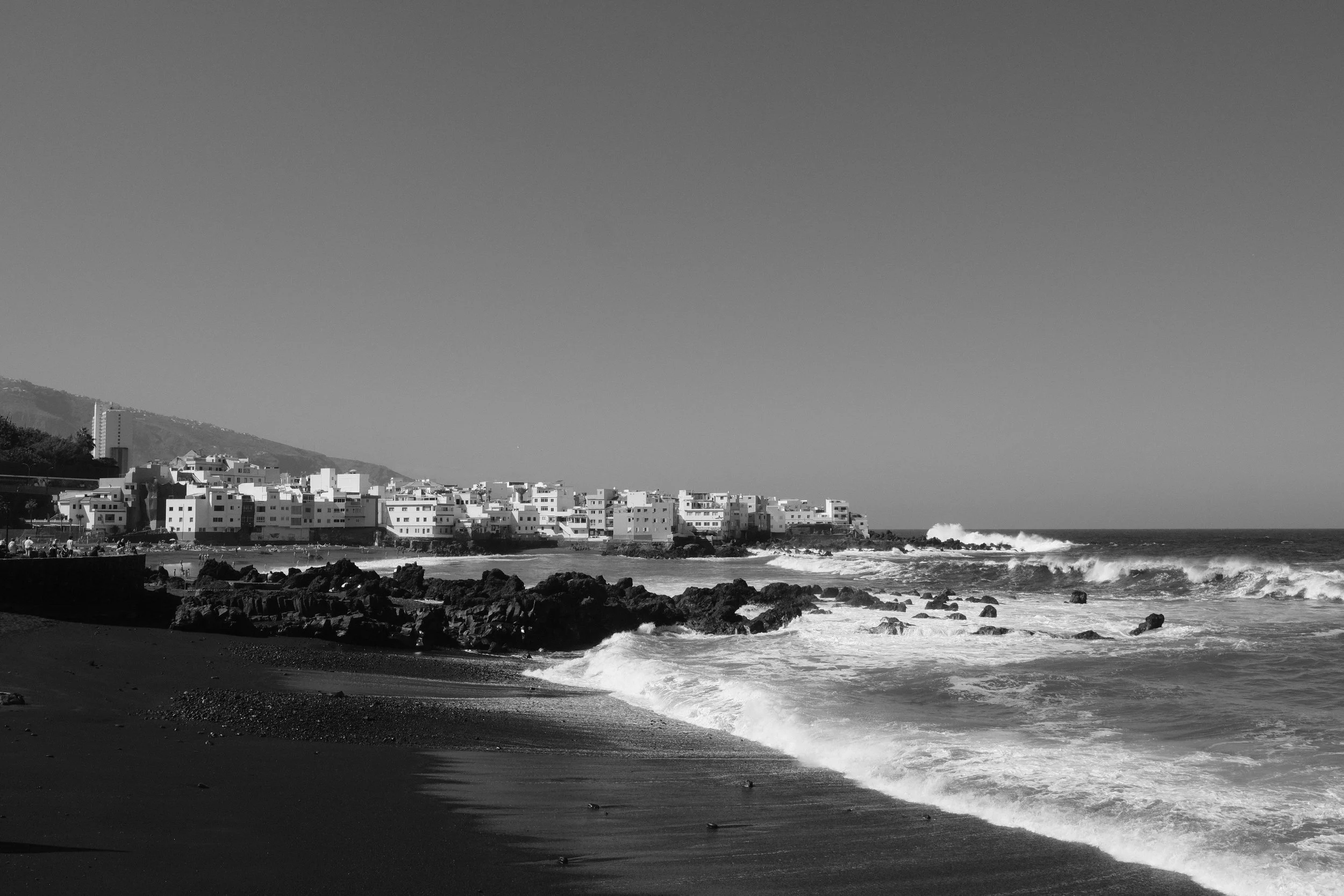 Schwarz-weiß-Foto eines Strands mit Felsen, Wellen und einem Stadtteil mit mehreren Gebäuden im Hintergrund