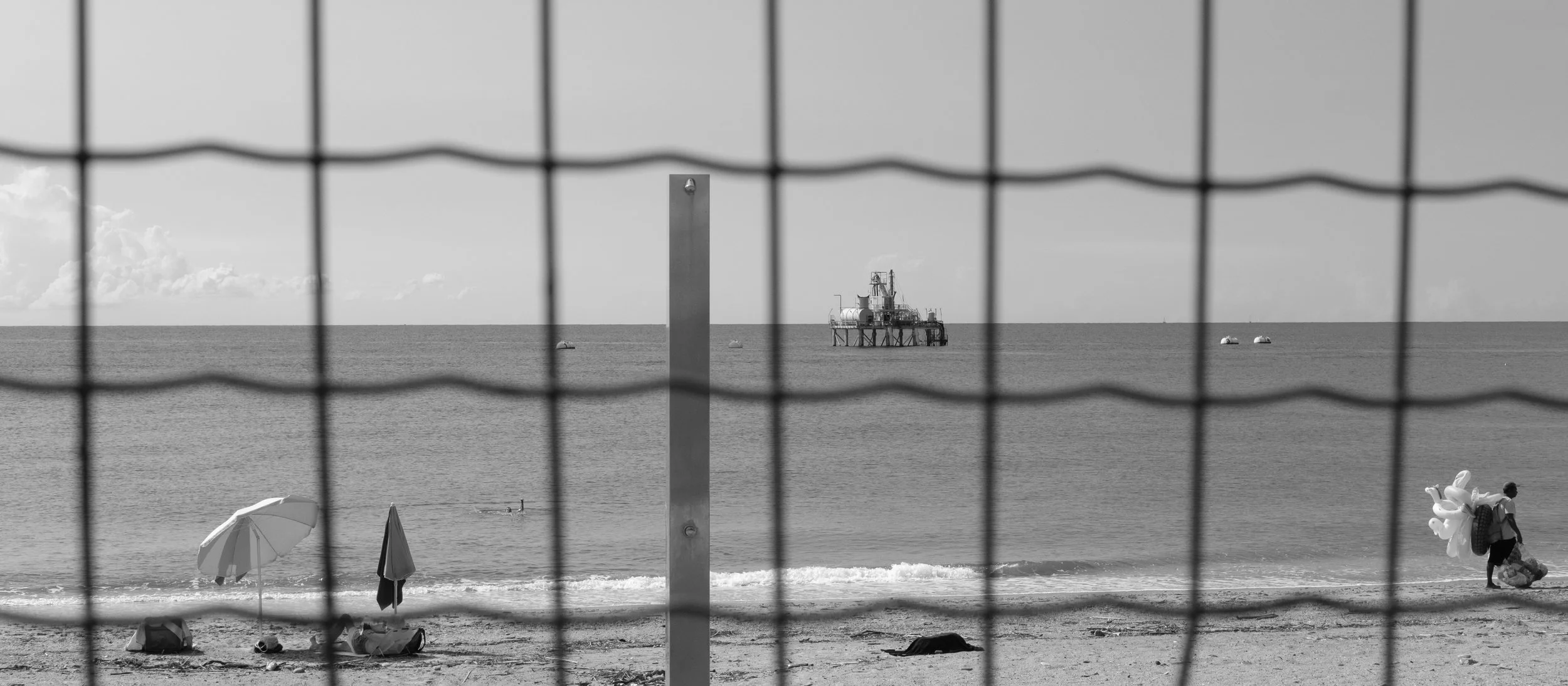 Strand mit Sonnenschirmen, eine Person mit Luftballons und Fischerboot im Hintergrund, Bild in Schwarz-Weiß, durch einen Drahtzaun betrachtet.