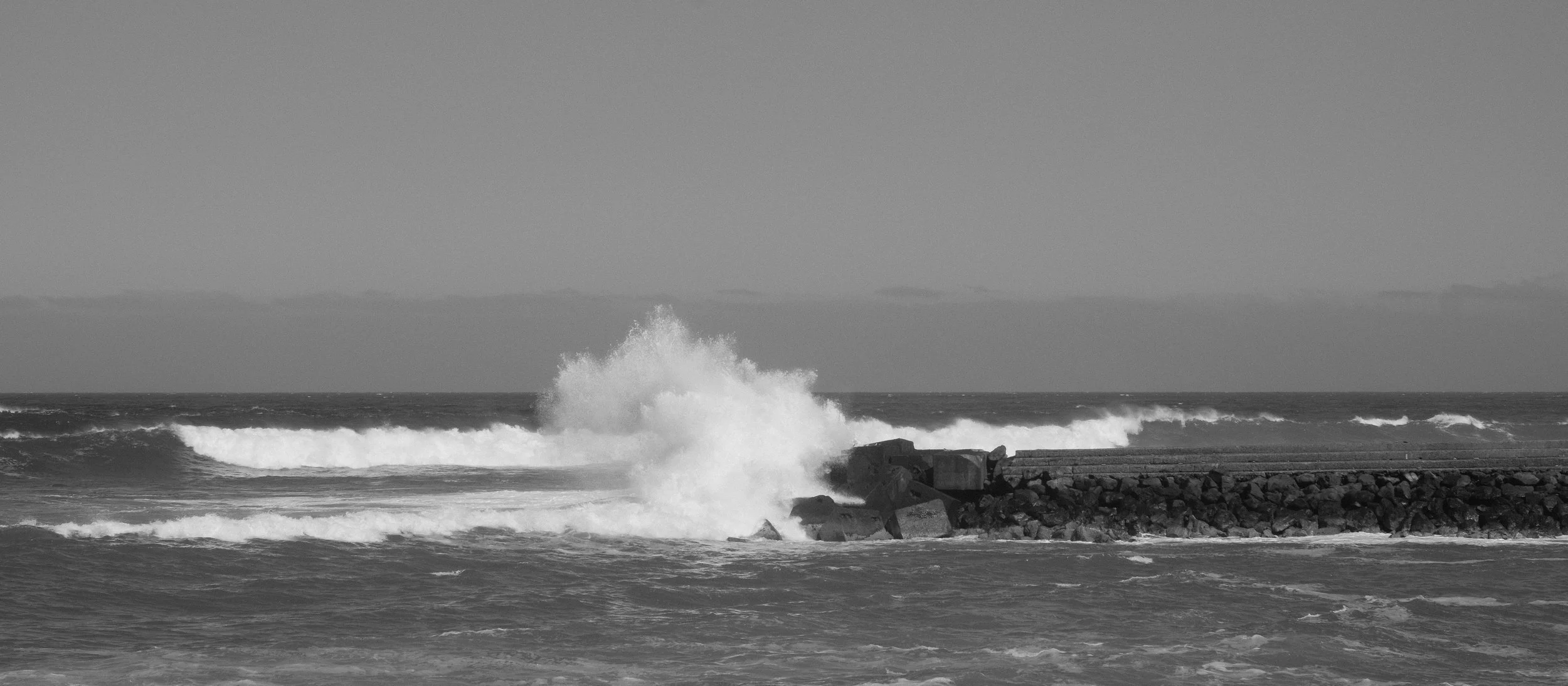Schwarzer und weißer Meeresküste mit brecherischen Wellen, die gegen eine Hafeneinfahrt mit Felsen schlagen, in Schwarz-Weiß.