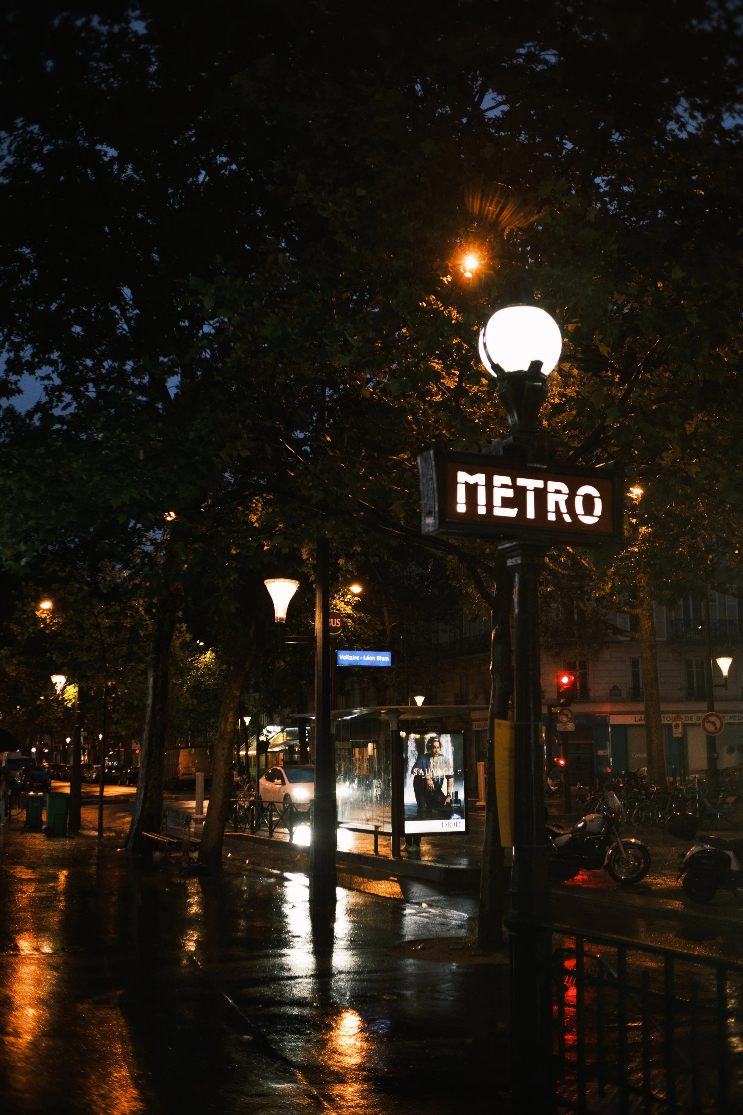 Straßenbahn Haltestelle bei Nacht mit beleuchtetem METRO Schild, Straßenlaternen, Bäumen und einer Werbetafel im Hintergrund.