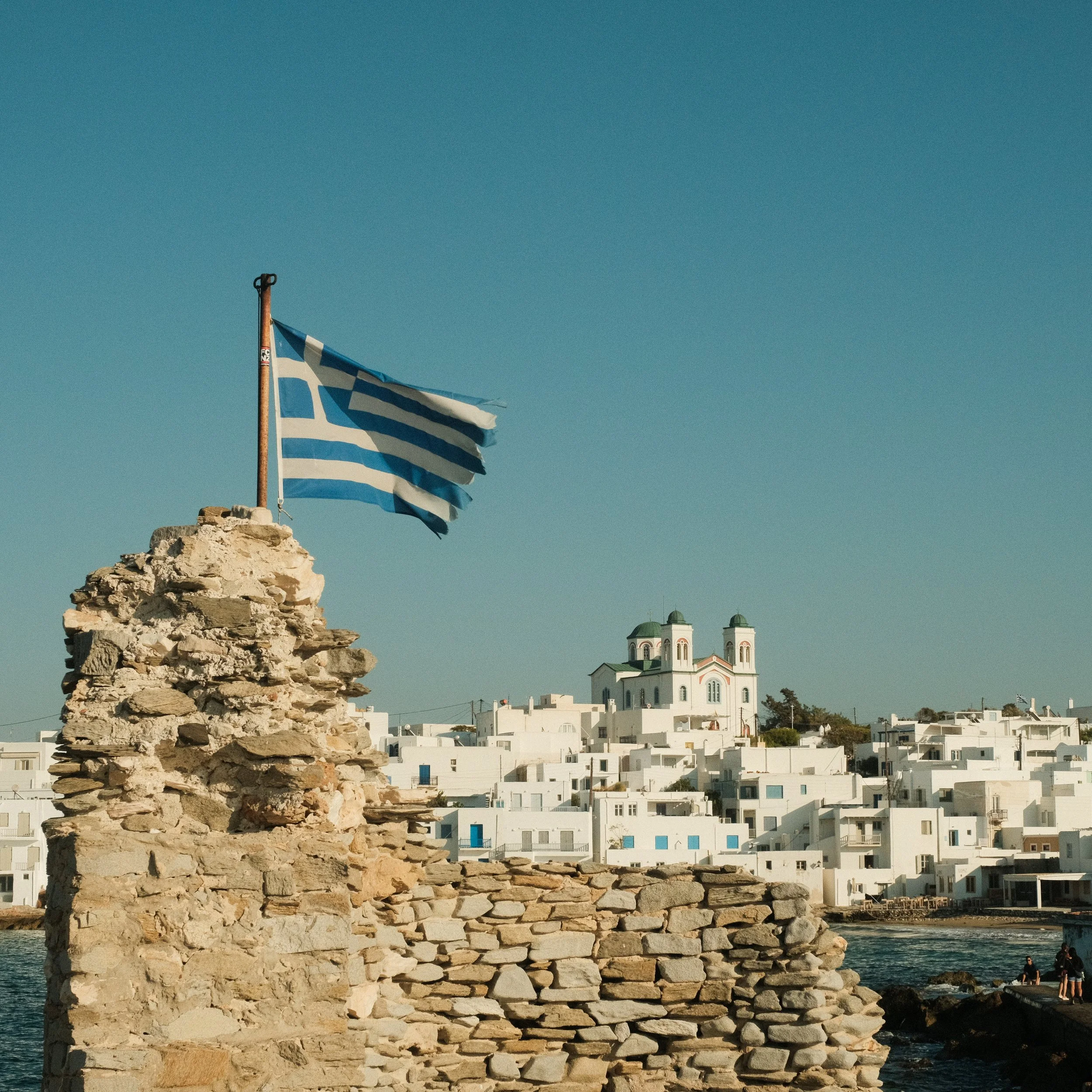Blick auf eine griechische Insel mit einer Flagge am Felsen im Vordergrund und weißen Häusern und einer Kirche im Hintergrund.