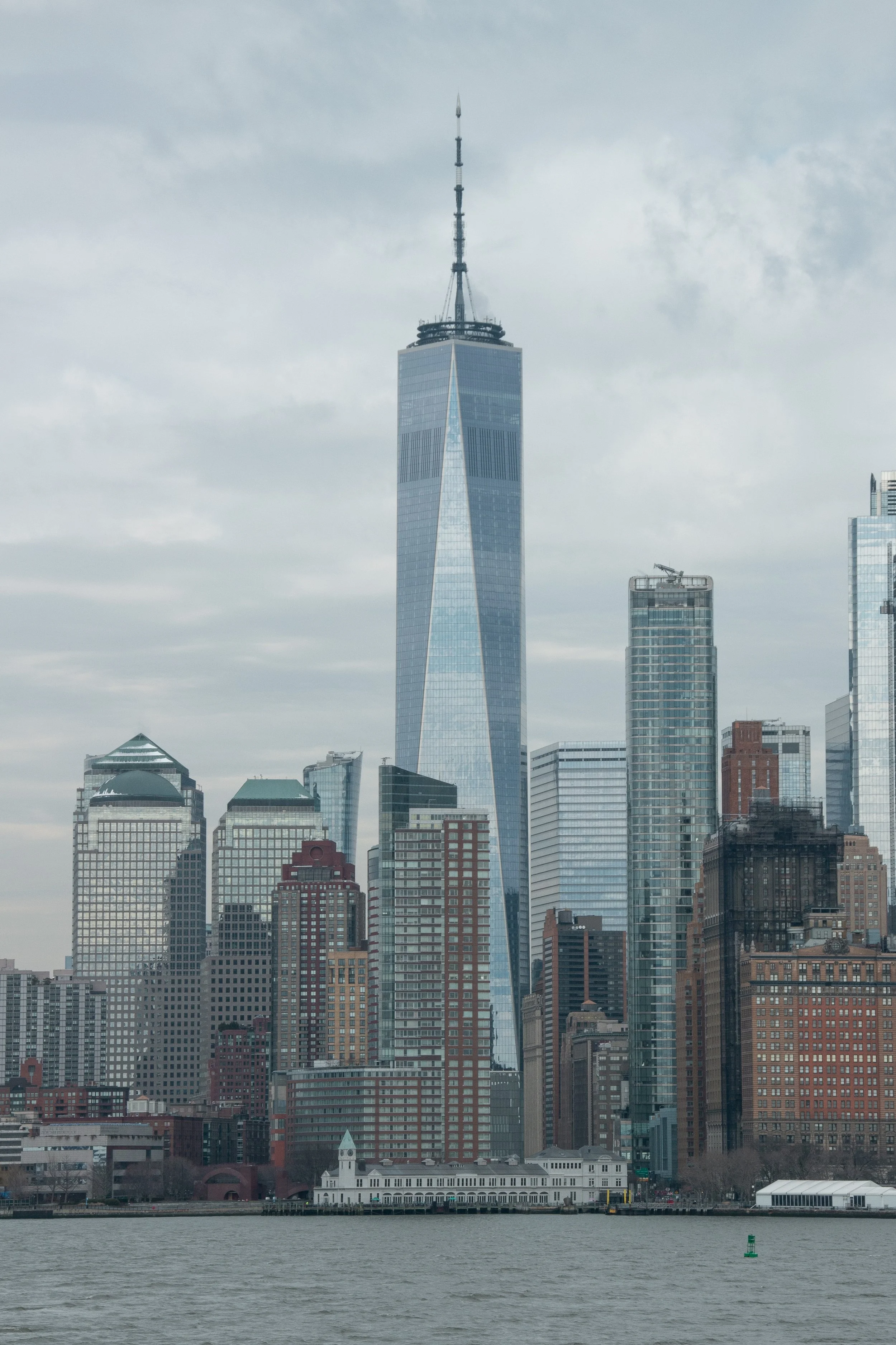 Stadtbild von New York mit One World Trade Center im Zentrum, aufgenommen vom Wasser aus bei bewölktem Himmel.