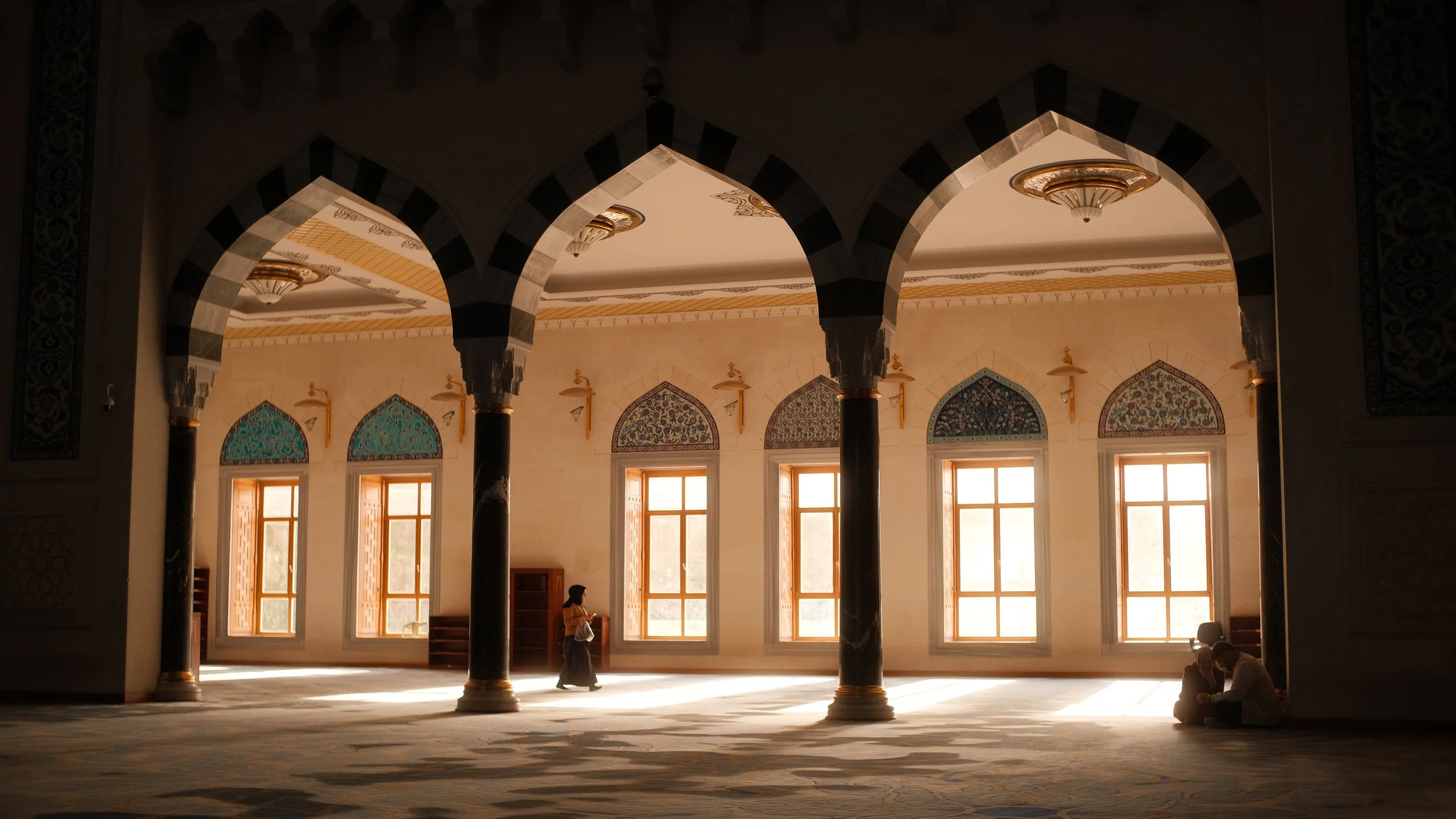 Innenraum eines islamischen Moschees mit hohen Bogenfenstern, schwarzen Säulen mit goldenen Details und hellen Wänden, durch die warmes Sonnenlicht scheint. Drei Menschen sind im Raum, zwei sitzen in einer Ecke, eine Person läuft.