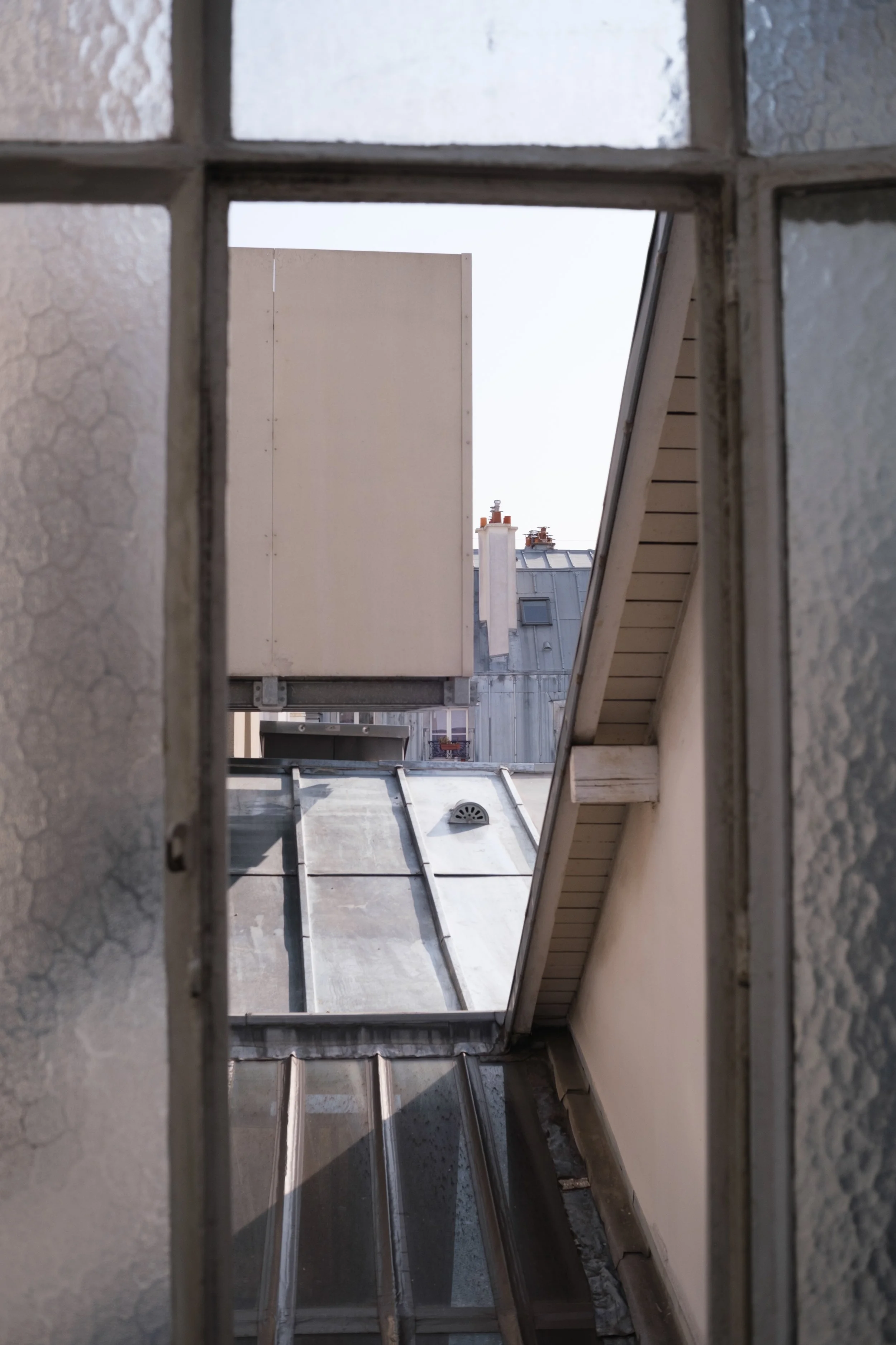 Blick durch ein kleines Fenster auf das Dach eines Hauses mit Metallblechen und Schornsteinen