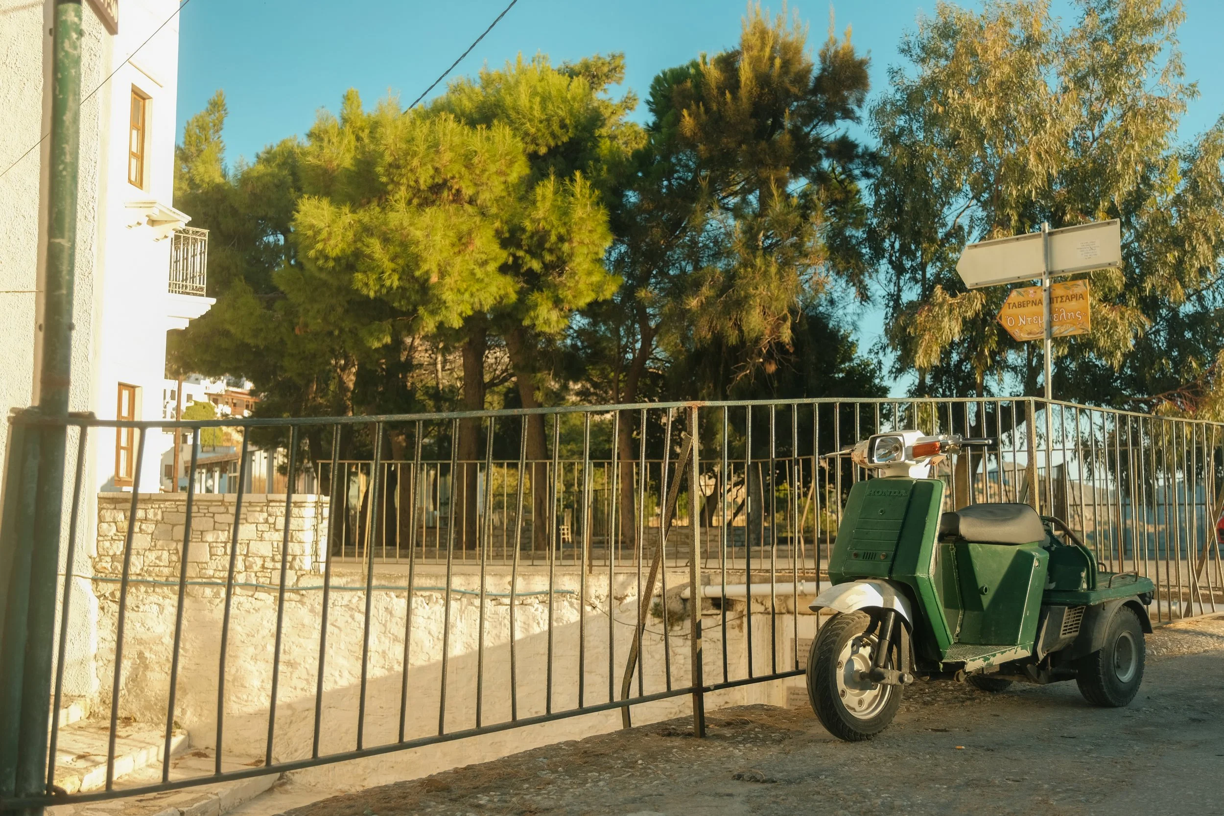 Ein grüner Motorroller steht auf dem Bürgersteig neben einer Metallbarriere, im Hintergrund sind Bäume und ein Gebäude mit Fenster und Balkon sichtbar.