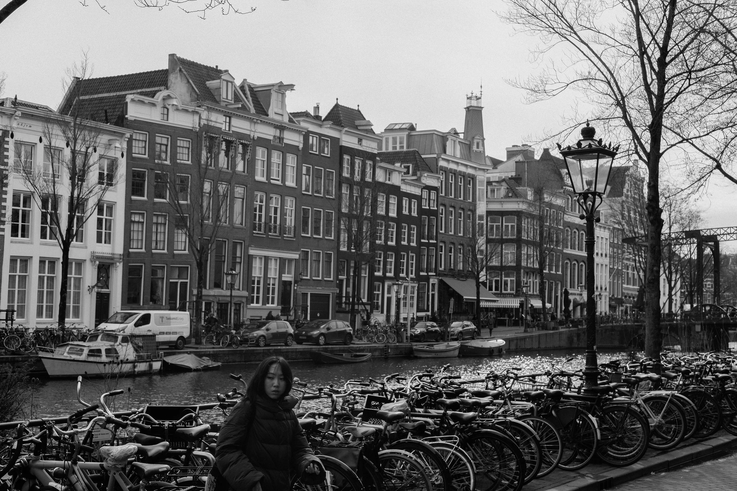 Ein Schwarz-Weiß-Foto einer Frau, die neben einem Fahrradständer an einem Kanal in Amsterdam steht, mit Häusern und Booten im Hintergrund.