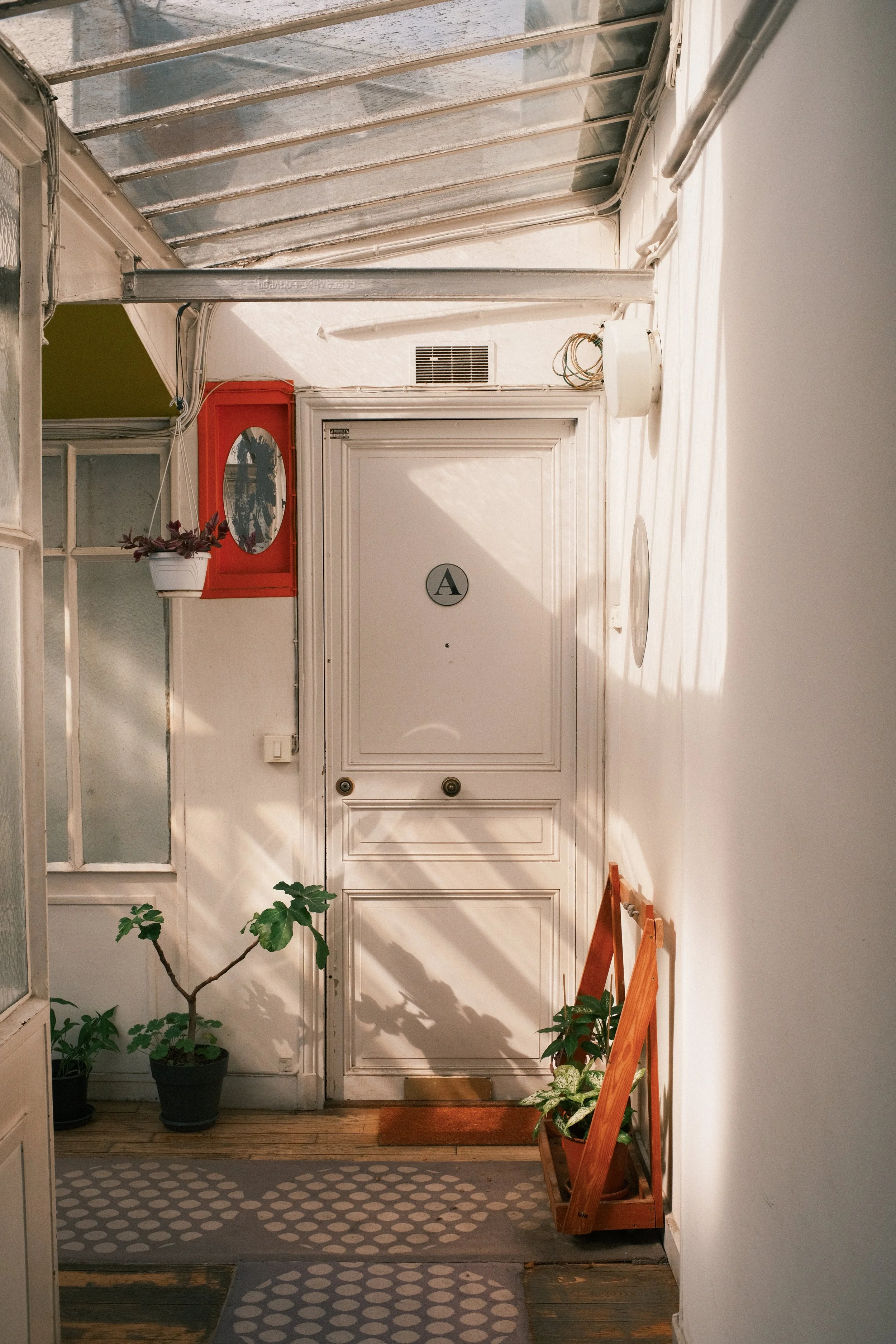 Weiße Tür mit dem Buchstaben 'A' in einem Kreis, im Flur mit Pflanzen, Polstermatten, einem roten Feuermelder und einem Kasten mit Spiegel.