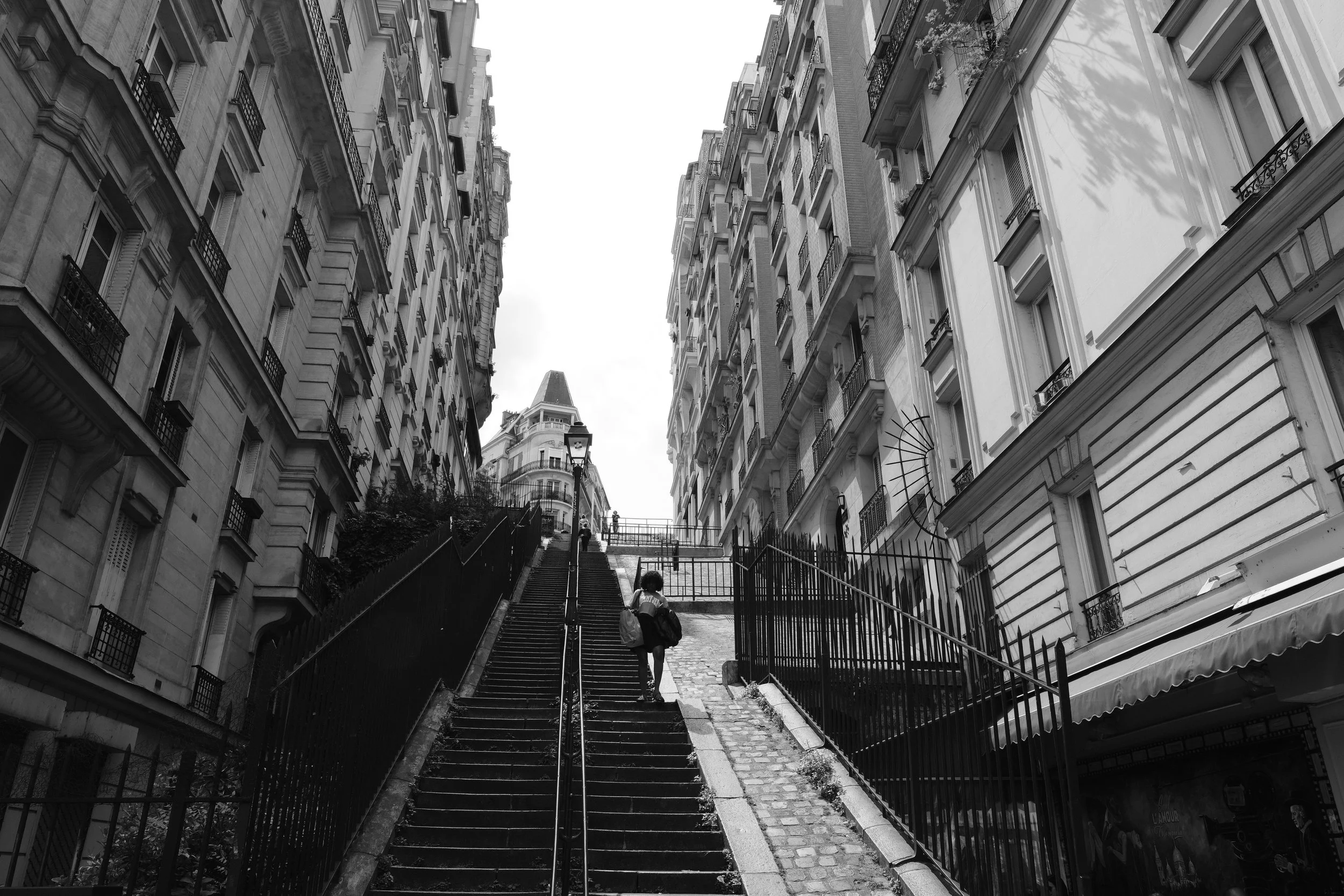 Schwarz-weiße Aufnahme einer steilen Treppe in einem städtischen Viertel mit hohen, ornamentalen Gebäuden auf beiden Seiten und einer Person, die die Treppe hinaufgeht.