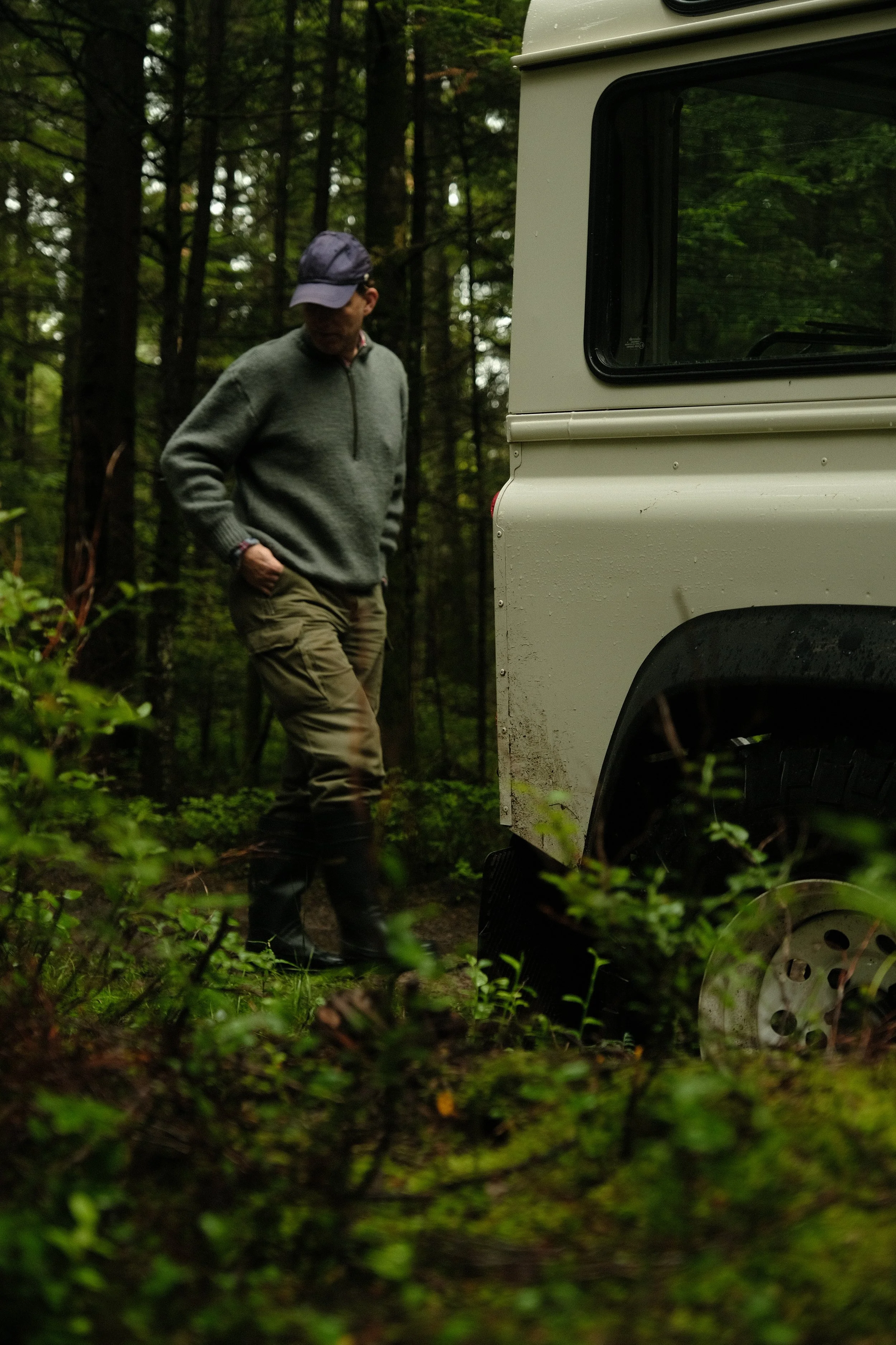 Ein Mann im Wald steht neben einem weißen Fahrzeug, trägt Outdoor-Kleidung und Gummistiefel.