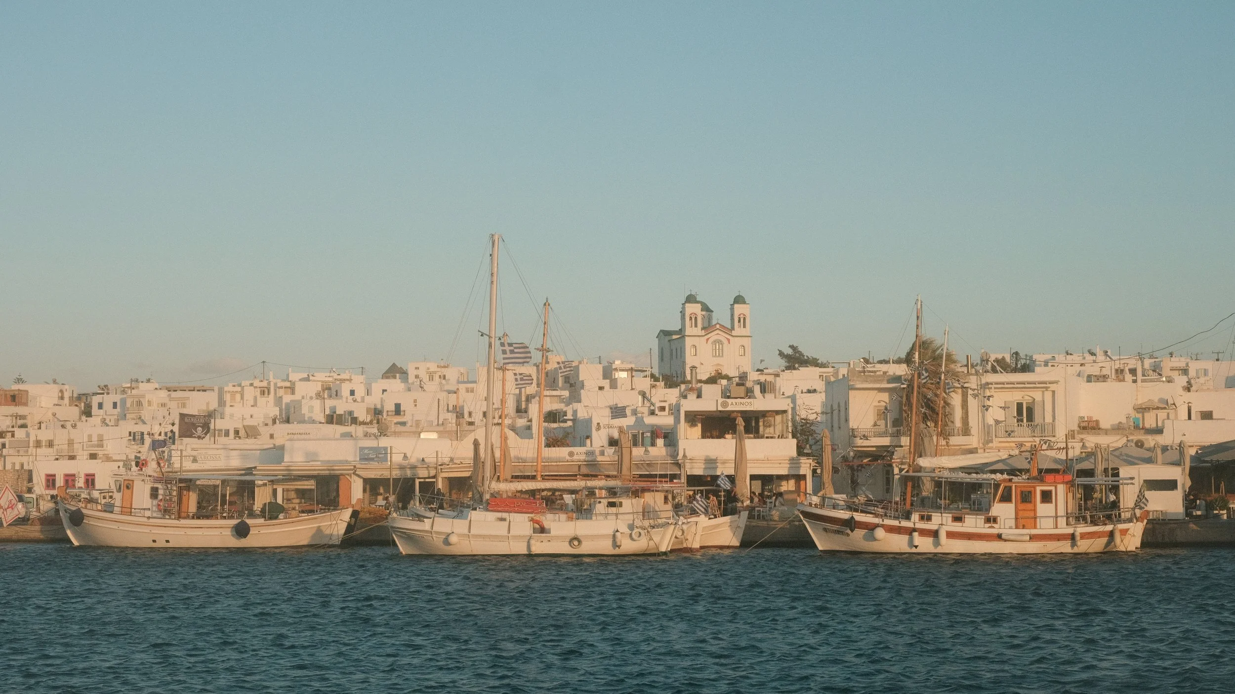 Blick auf einen Hafen mit drei Booten und weißen Gebäuden im Hintergrund, inklusive einer Kirche mit zwei Türmen.