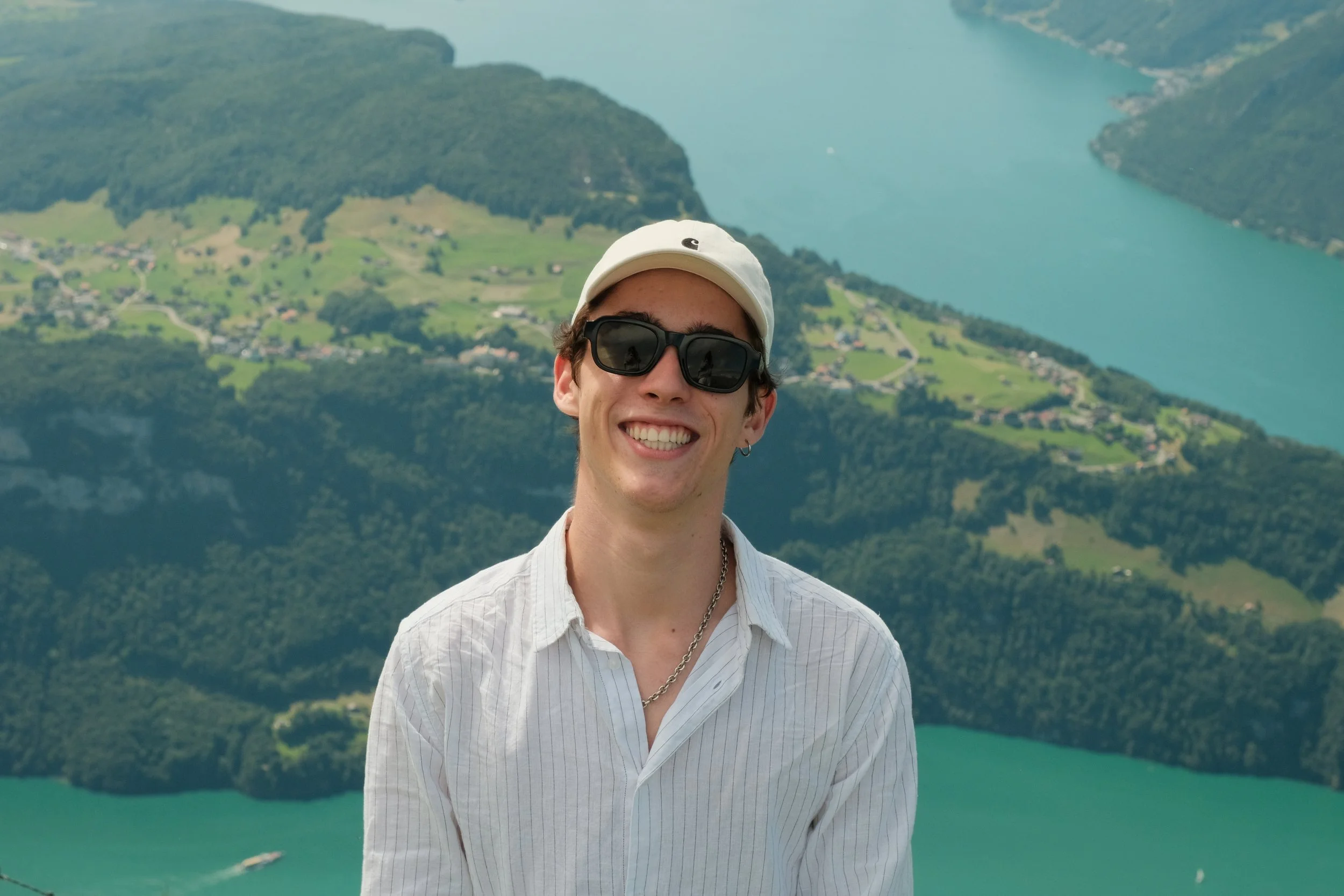 Ein junger Mann mit Sonnenbrille, weißen Hemd, weißer Kappe und Silberkette steht vor einer malerischen Seenlandschaft mit Hügeln und Wasser im Hintergrund.