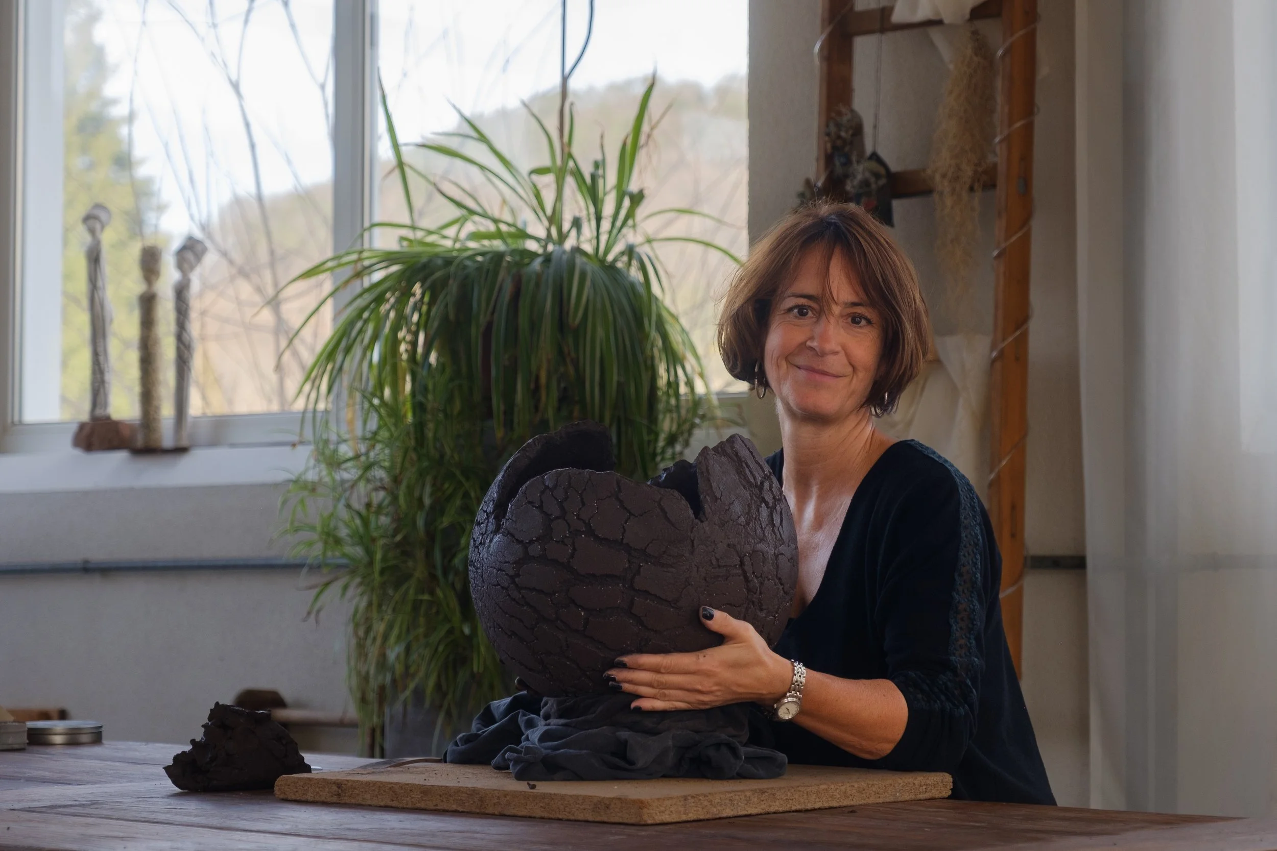 Eine Frau sitzt an einem Tisch und hält eine große, geformte Skulptur aus dunklem Material in den Händen. Im Hintergrund sind große Fenster, durch die Natur mit Bäumen zu sehen ist, und drinnen sind einige Pflanzen und Dekorationen.
