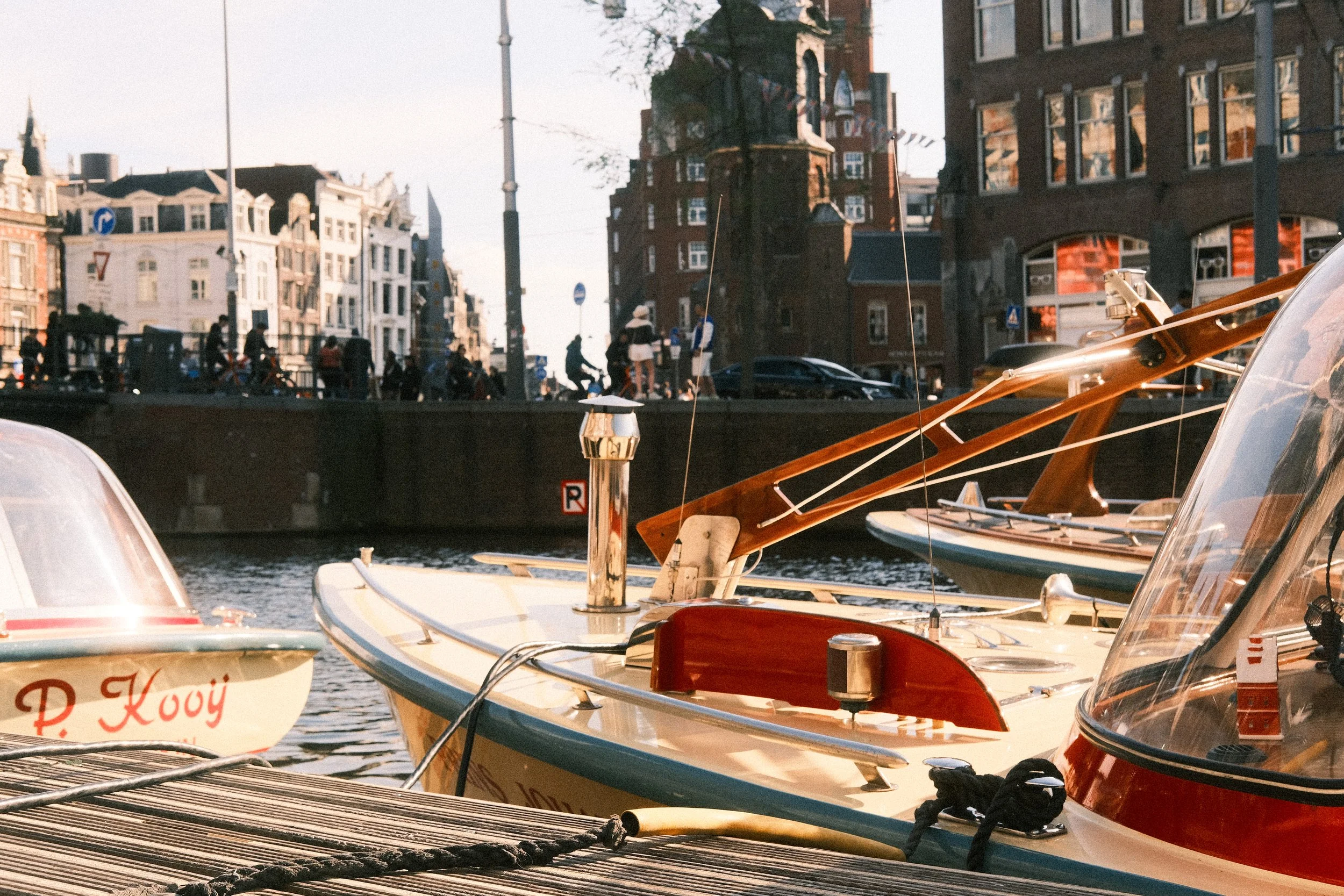 Boote liegen am Dock in Amsterdam mit Blick auf eine Stadt mit historischen Gebäuden und Brücken, im Sonnenlicht.