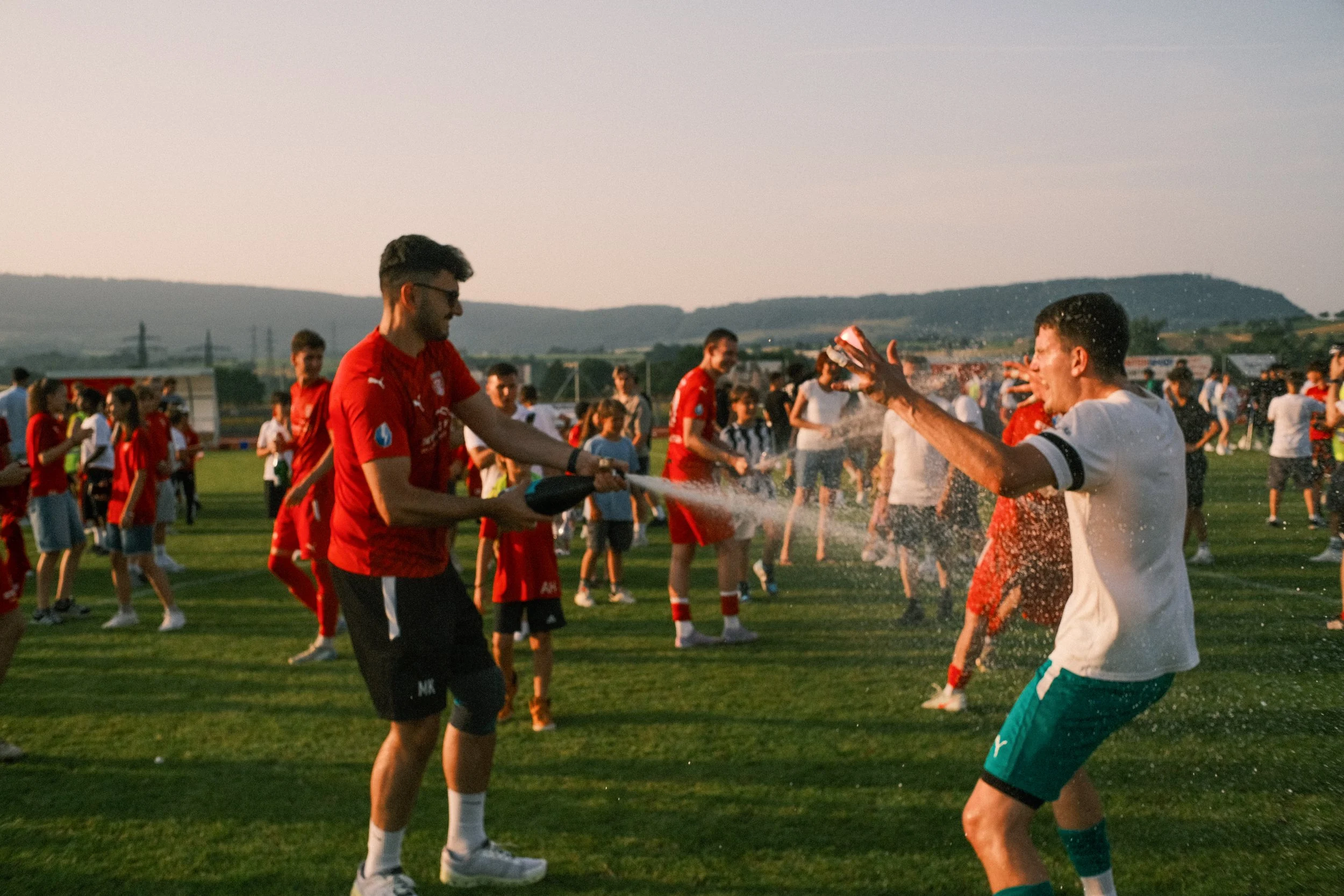 Fußballspieler feiern auf einem Rasenplatz, einer sprüht Sekt in die Luft, während die anderen jubeln und Wasser sprüht, im Hintergrund sind mehr Menschen und Berge bei Sonnenuntergang.