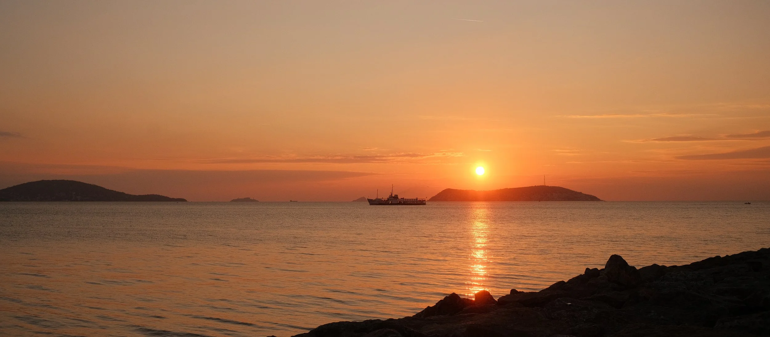 Ein Sonnenuntergang über dem Meer mit einem Schiff in der Mitte und Inseln im Hintergrund, ruhiges Wasser und dunkler Küstenstreifen im Vordergrund.