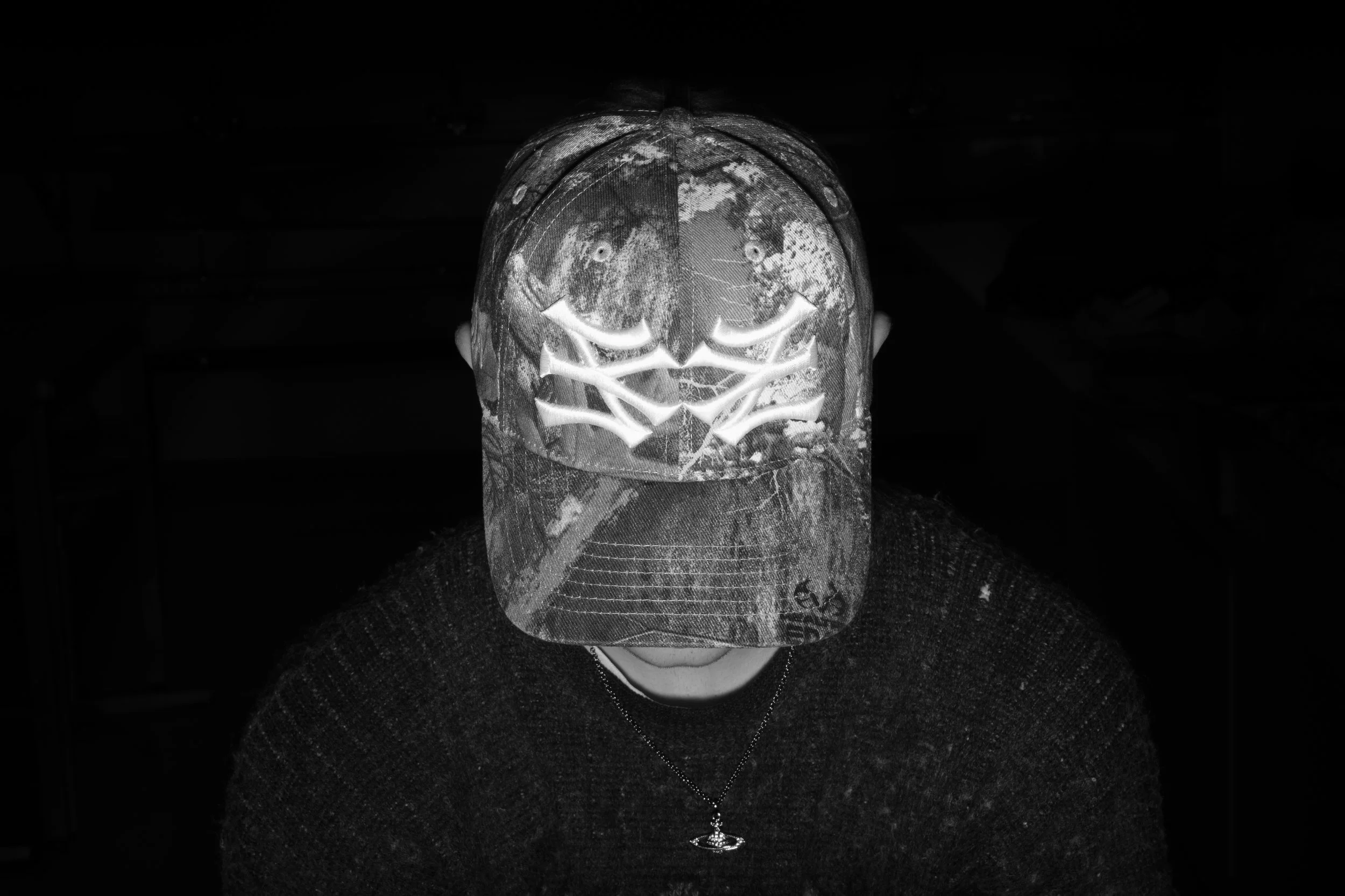 Mann trägt eine Camouflage-Kappe mit sichtbarem Totenkopf-Design und eine Halskette mit einem Planetenanhänger, blickt nach unten, vor einem dunklen Hintergrund.