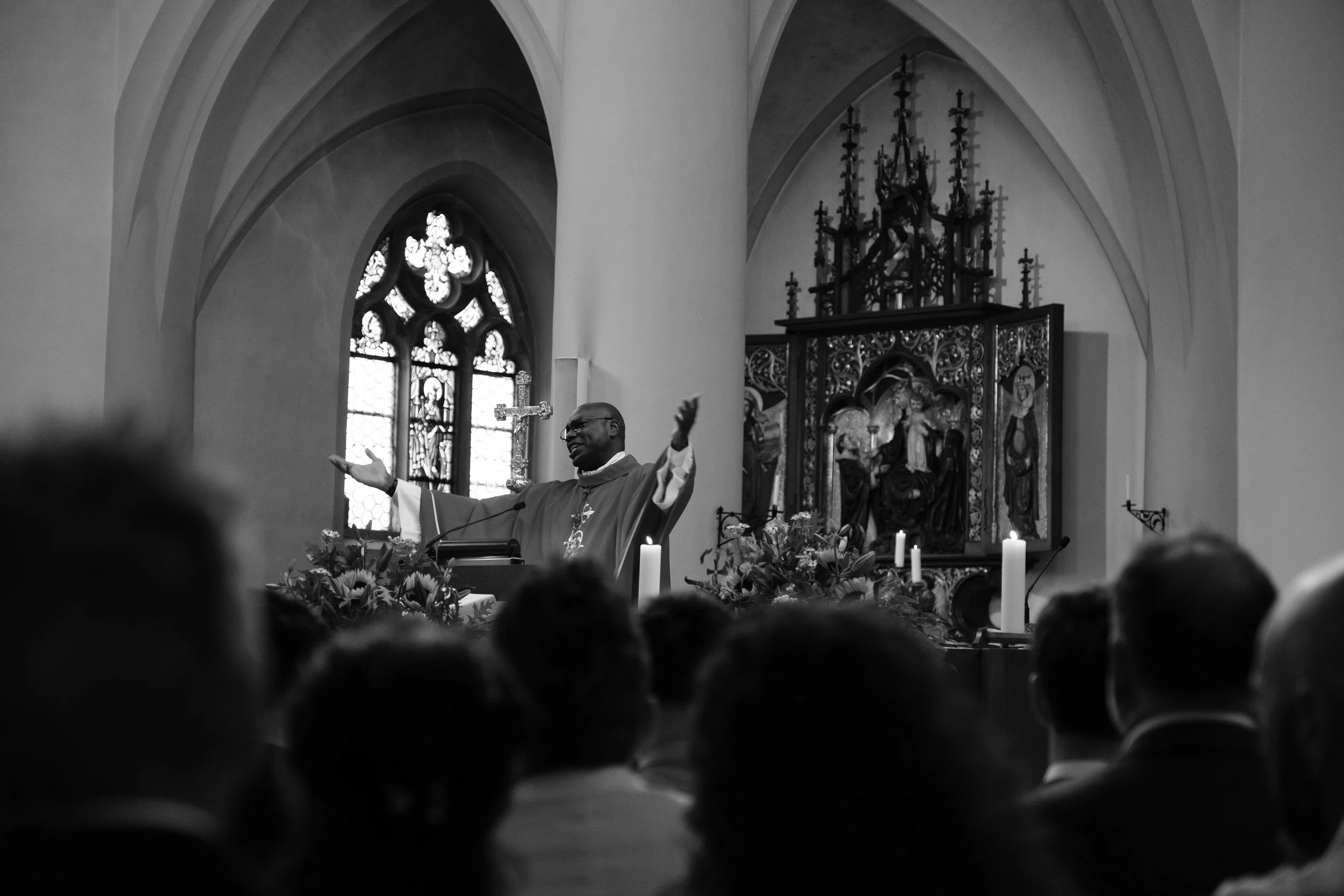 Ein Priester in der Kirche, der mit ausgestreckten Armen spricht, während die Gemeinde zuhört. Im Hintergrund sind Kirchenfenster, eine Kreuz-Statue und ein kunstvolles Altargemälde mit Kerzen.
