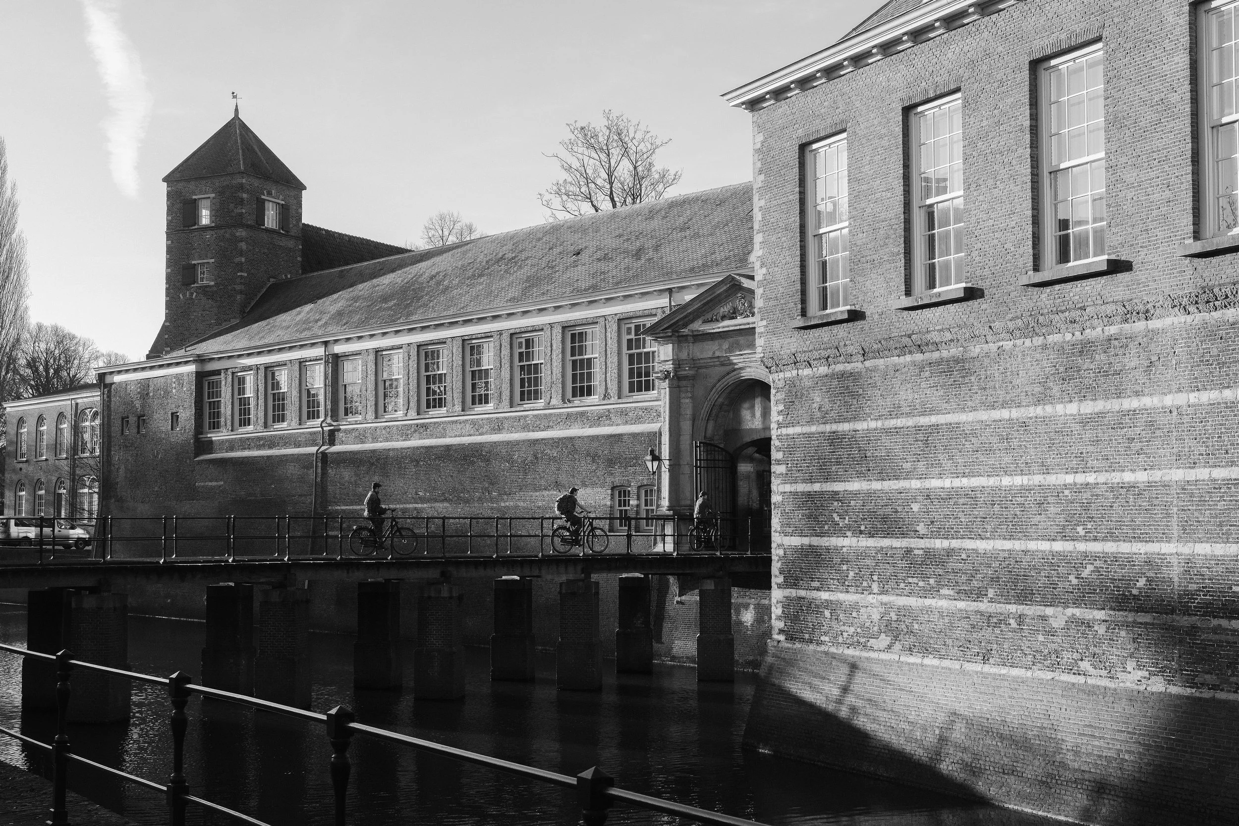 Schwarz-Weiß-Foto von einer alten Festung oder Burg mit Wasser im Vordergrund, einem Steg mit Fahrrädern und einem Tor als Eingang, im Hintergrund Bäume und Wolken am Himmel.