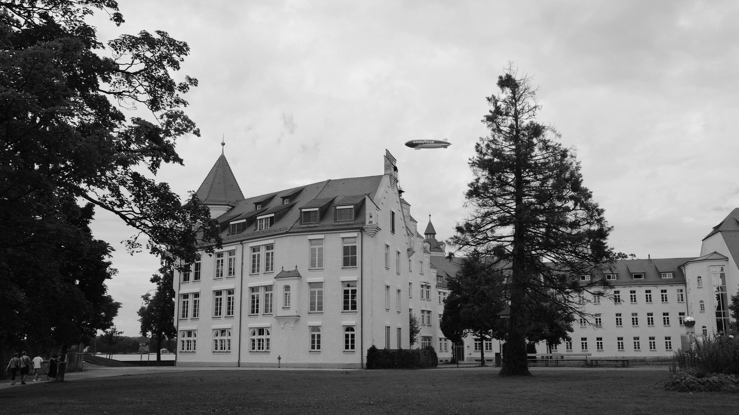 Ein großes, weißes Schloss mit mehreren Dachgauben, Türmen und vielen Fenstern, umgeben von Bäumen, mit einem Zeppelin im Himmel.