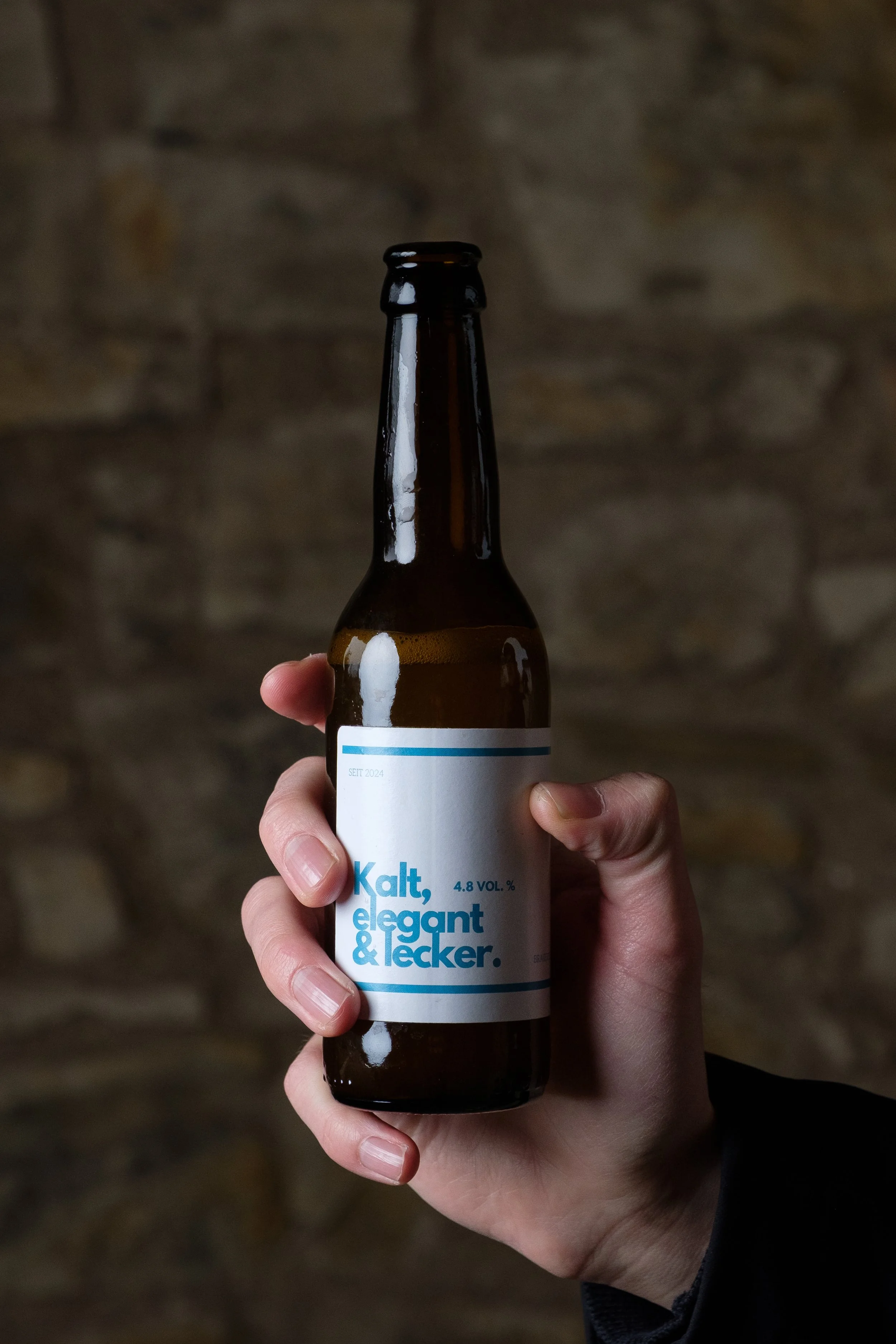 Eine Hand hält eine braune Bierflasche vor einem Steinmauerhintergrund, das Etikett ist weiß mit blauer Schrift, die Wörter sind 'Kalt, elegant & lecker'.