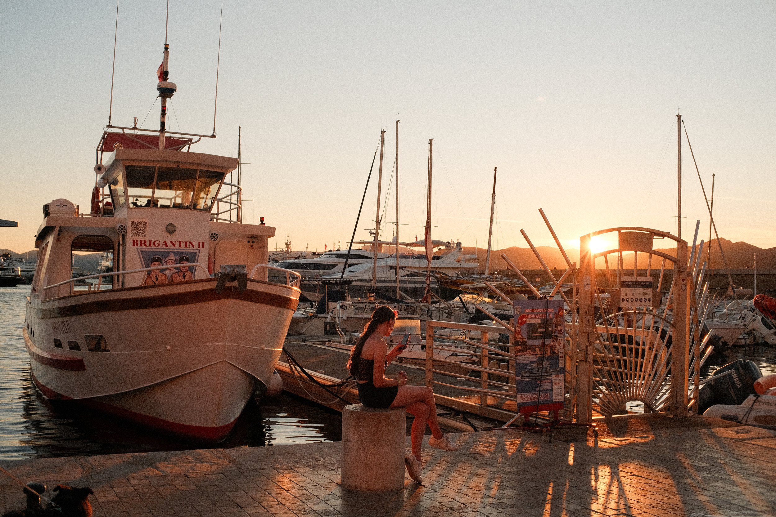 Eine junge Frau sitzt auf einer Betonmauer am Hafen, während die Sonne untergeht und Boote im Wasser liegen.
