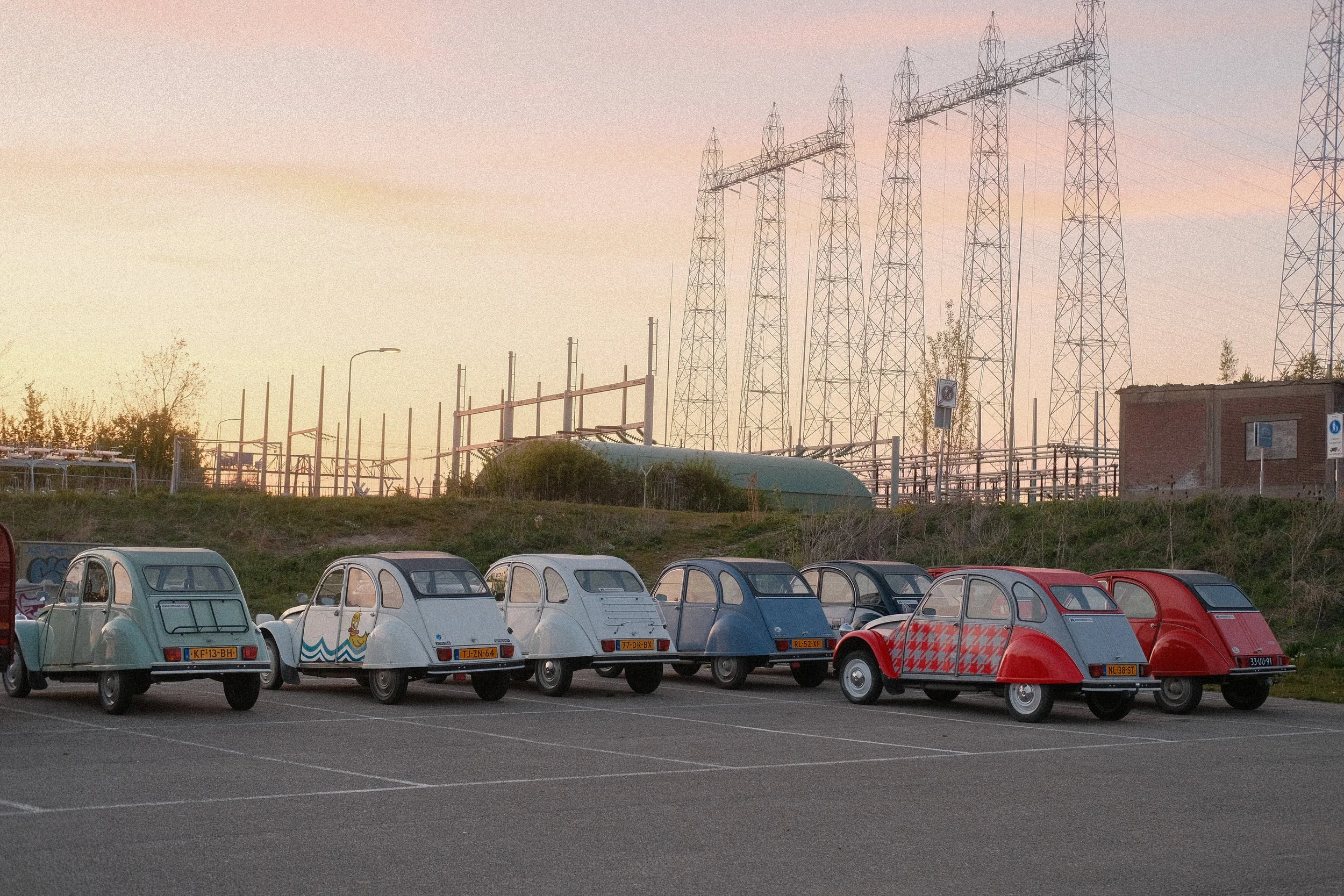 Mehrere kleine, oldschool Fahrzeuge in verschiedenen Farben, geparkt auf einem Parkplatz bei Sonnenuntergang, im Hintergrund Hochspannungsleitungen.