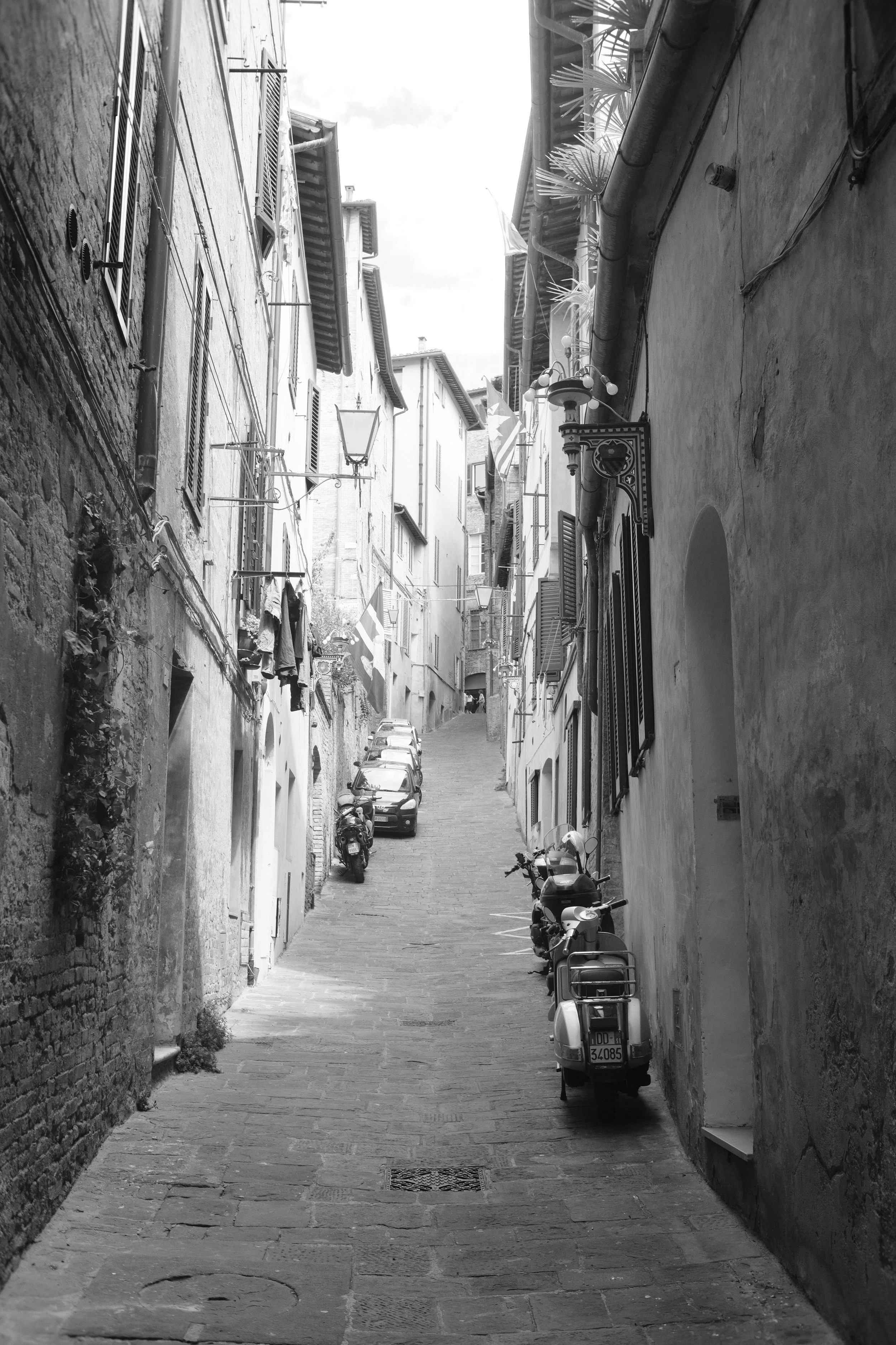 Schmale, steile Straße in einer europäischen Stadt mit Parkszenen, engen Häusern, Moped und Autos, im Schwarz-Weiß-Stil.