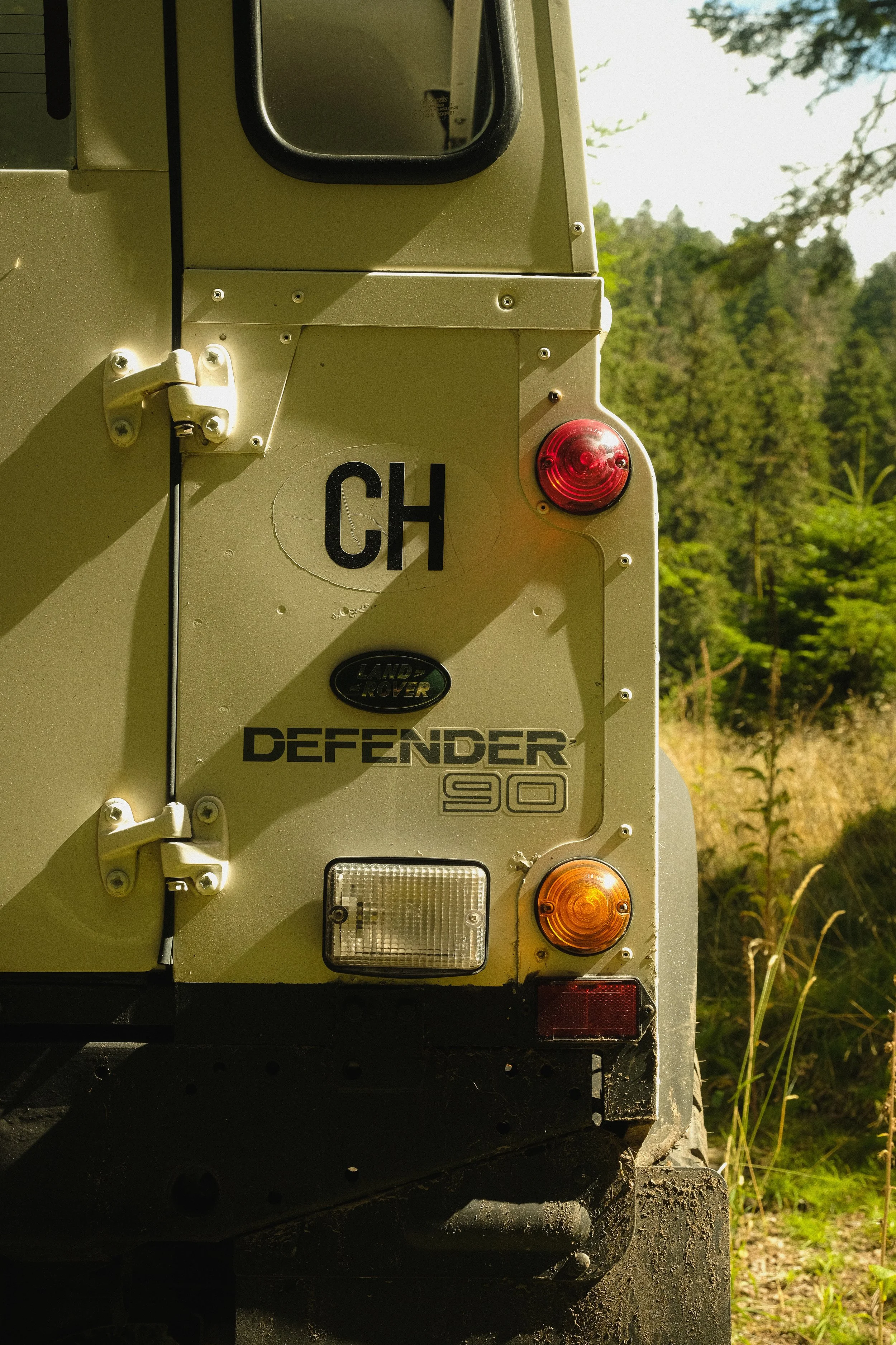 Rückseite eines Land Rover Defender 90, desertfarben, mit Scheinwerfern, Rücklichtern, Kennzeichen 'CH' und Markierungen, im Freien mit Bäumen im Hintergrund.