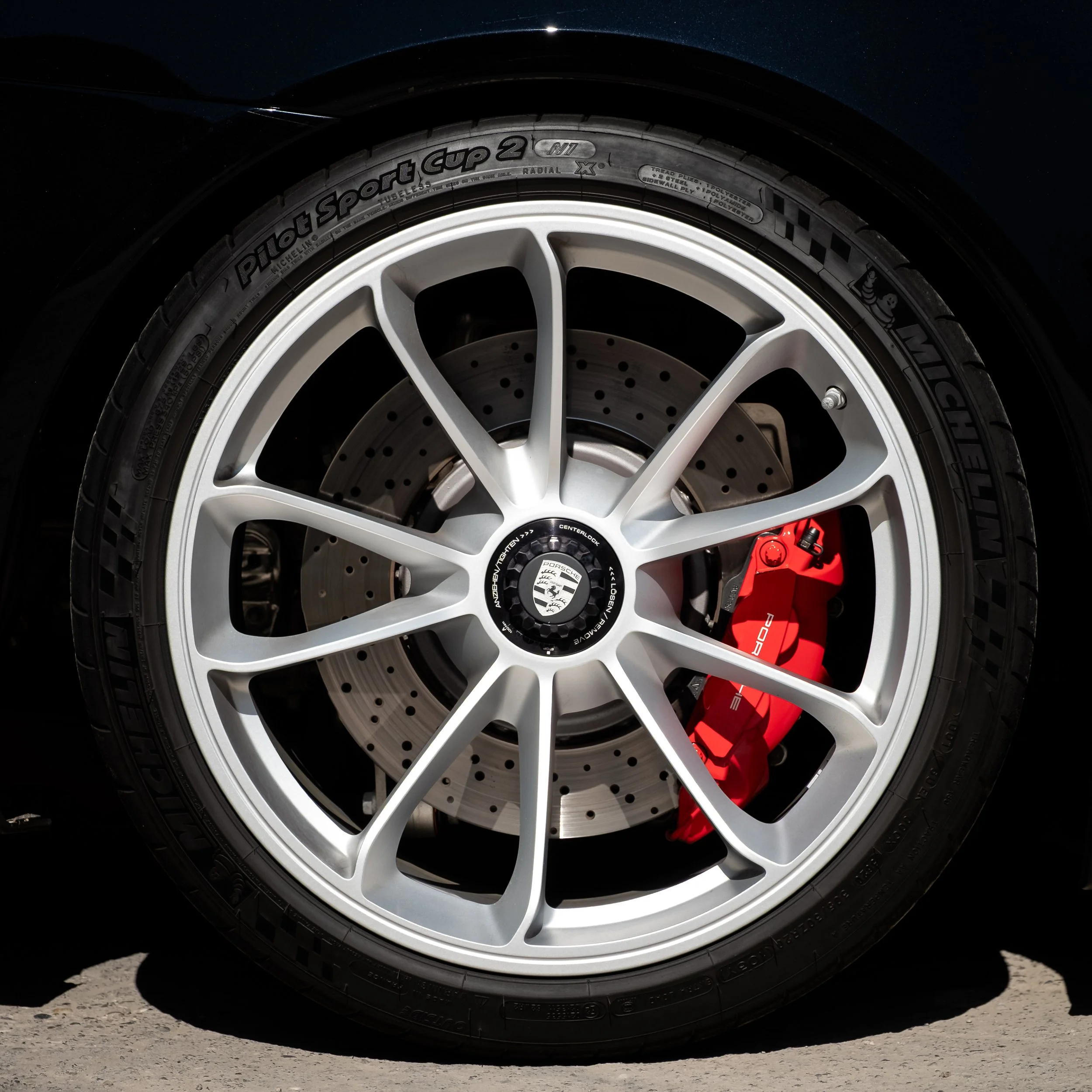 Nahaufnahme eines Auto-Reifen mit Sportfelge und breiter Bremsscheibe, roter Bremssattel mit Porsche-Logo.